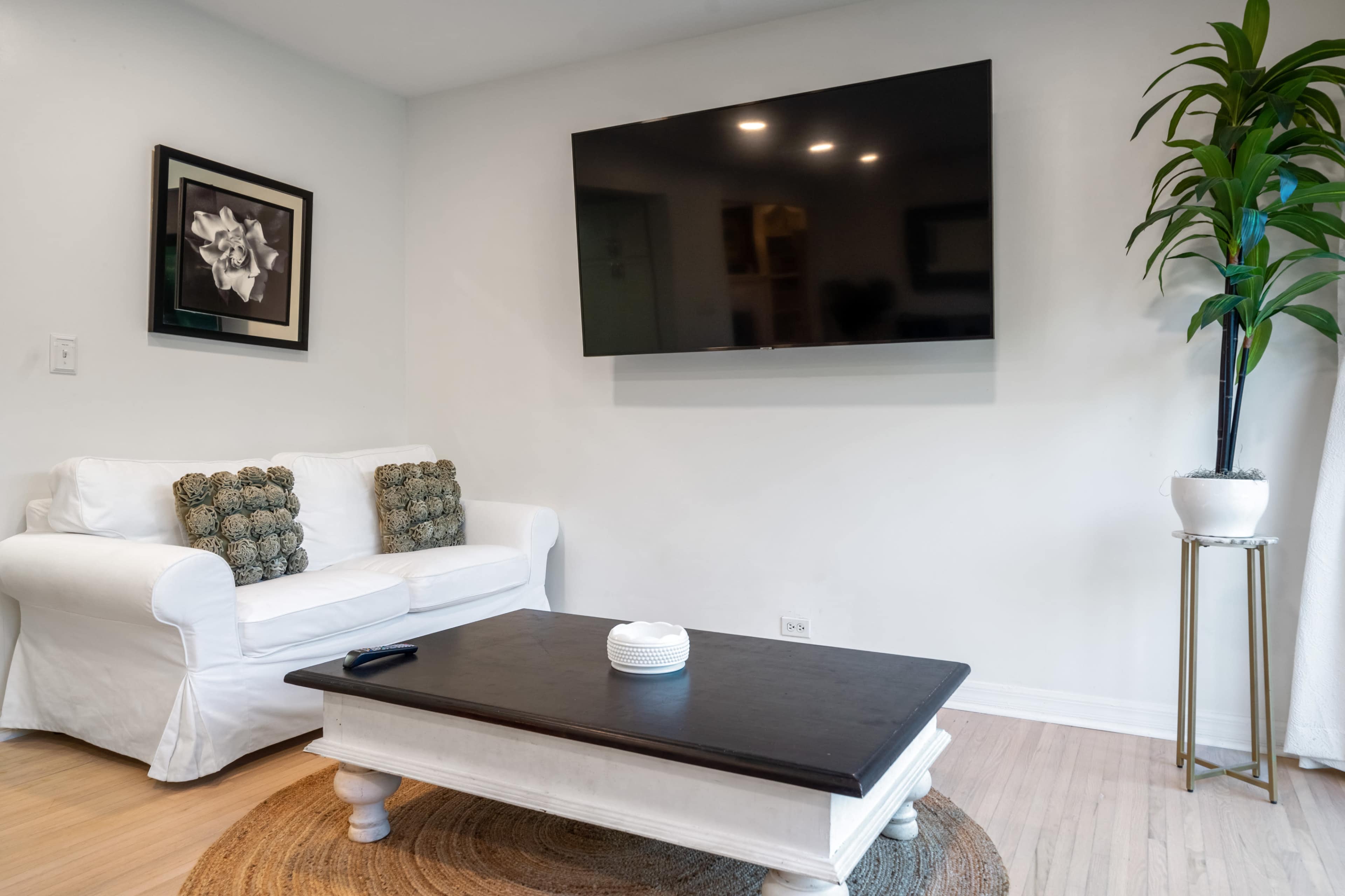 The living room features a white sofa with decorative pillows, a dark wood coffee table on a circular rug, and a wall-mounted television above the table.