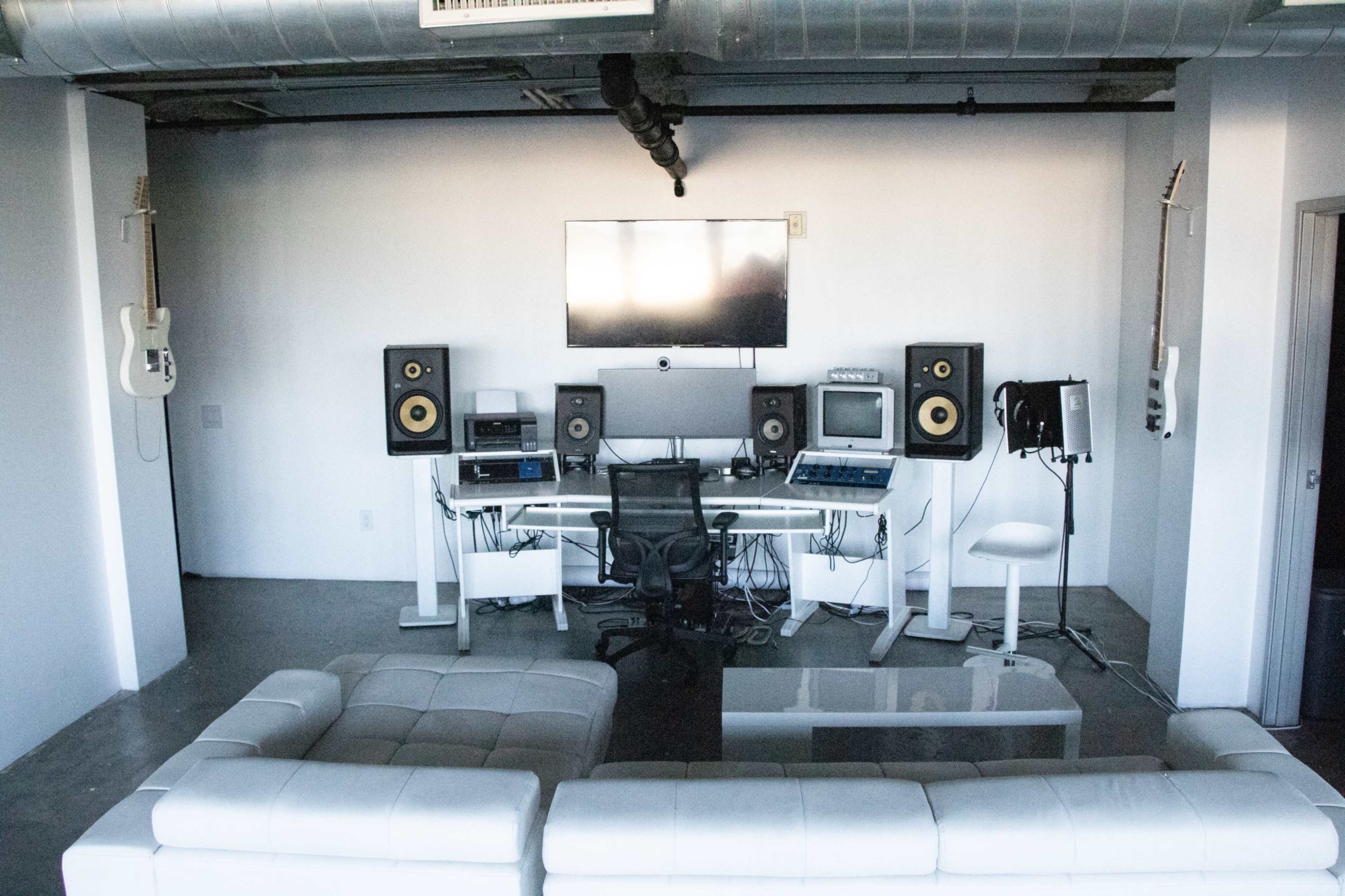 A modern music studio featuring a computer setup with audio equipment, speakers, and a large screen, accompanied by a white sectional sofa in the foreground.