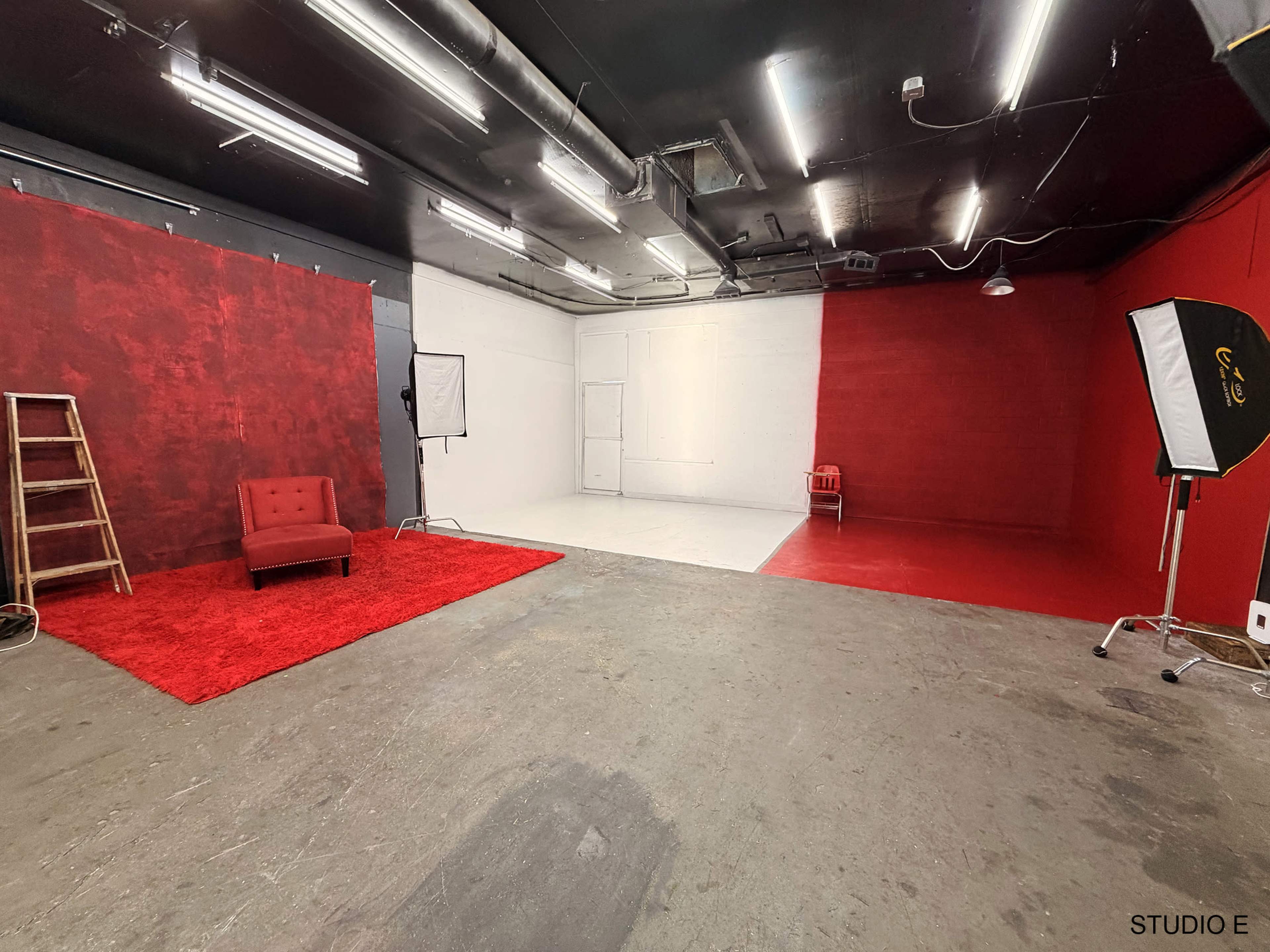 The image shows a photography studio divided into two sections: one side has a red backdrop with a red chair and rug, while the other side is plain white with a red wall.