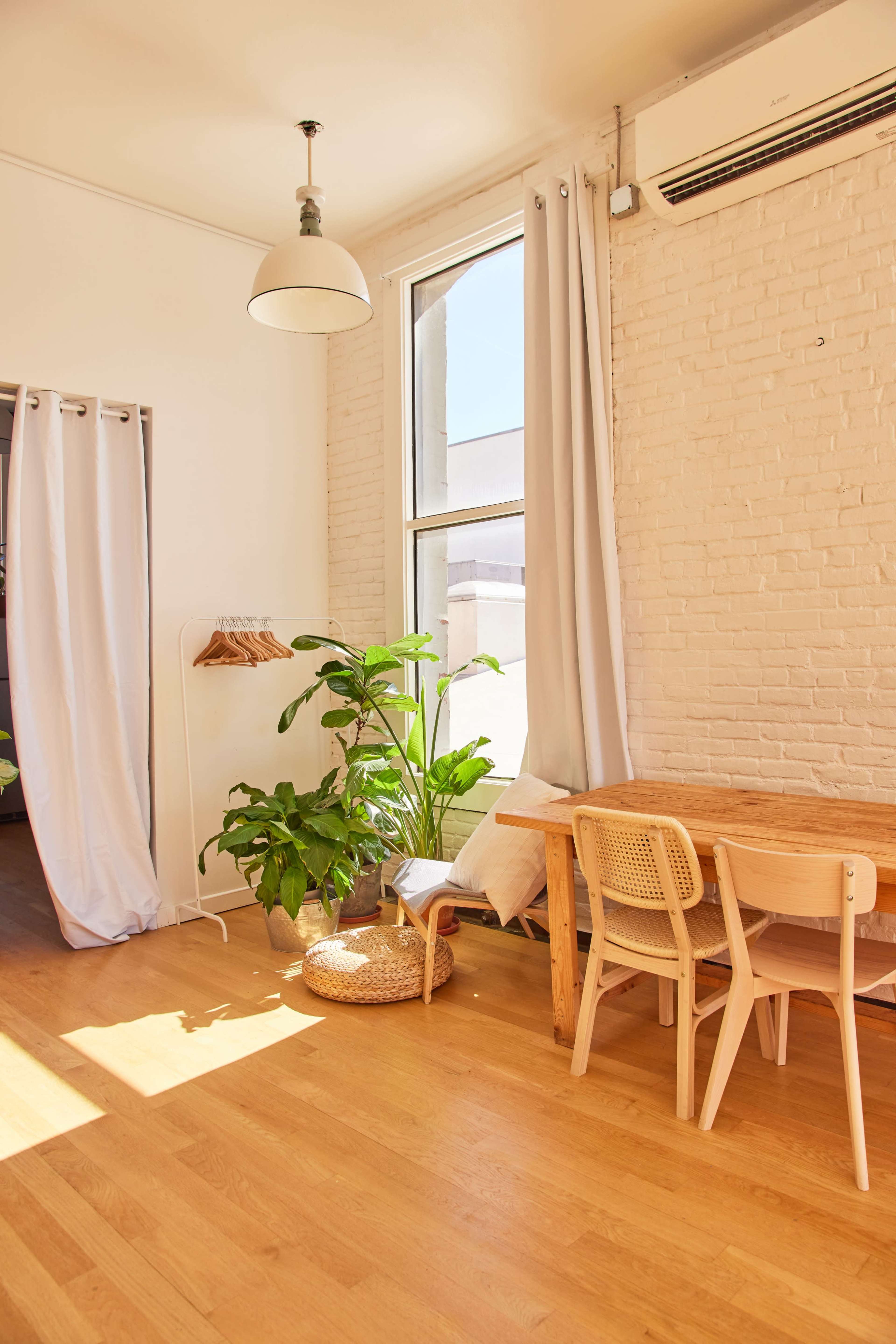 A bright, sunlit room features a wooden table and chairs, a potted plant, and light-colored curtains hanging by a large window.
