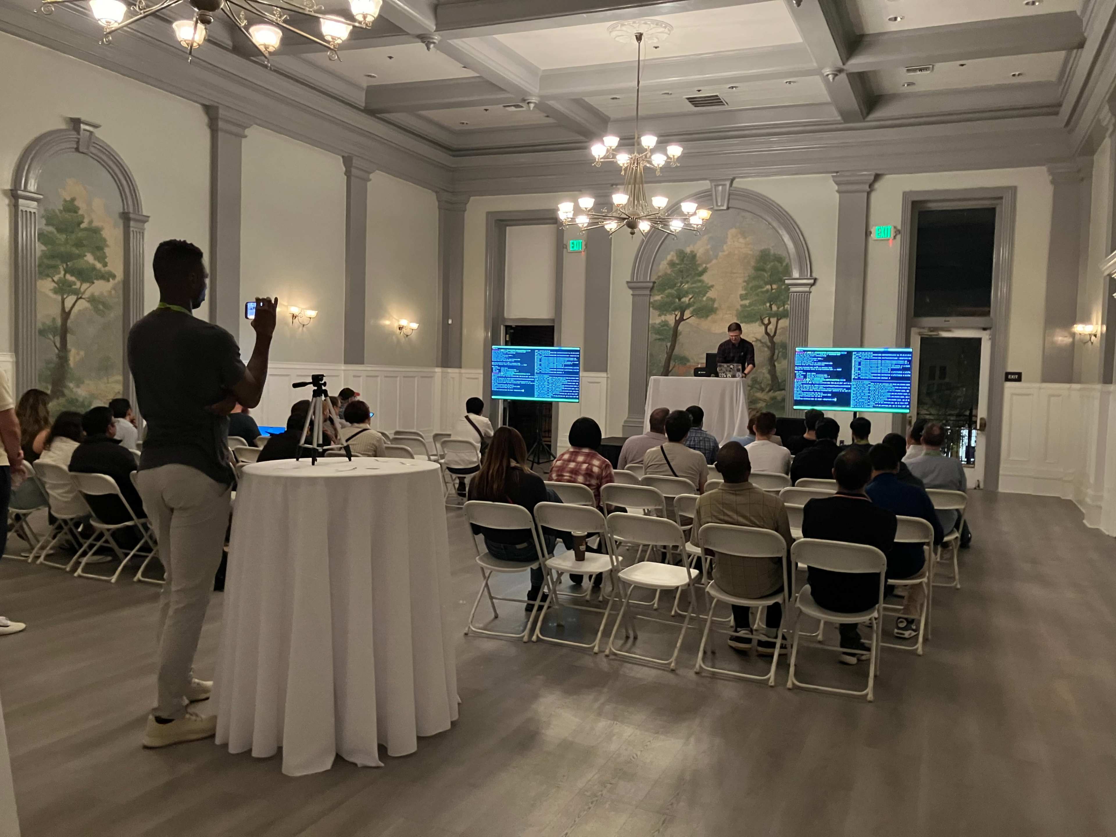 A speaker presents to an audience seated in a large room with decorative walls and two screens displaying information.