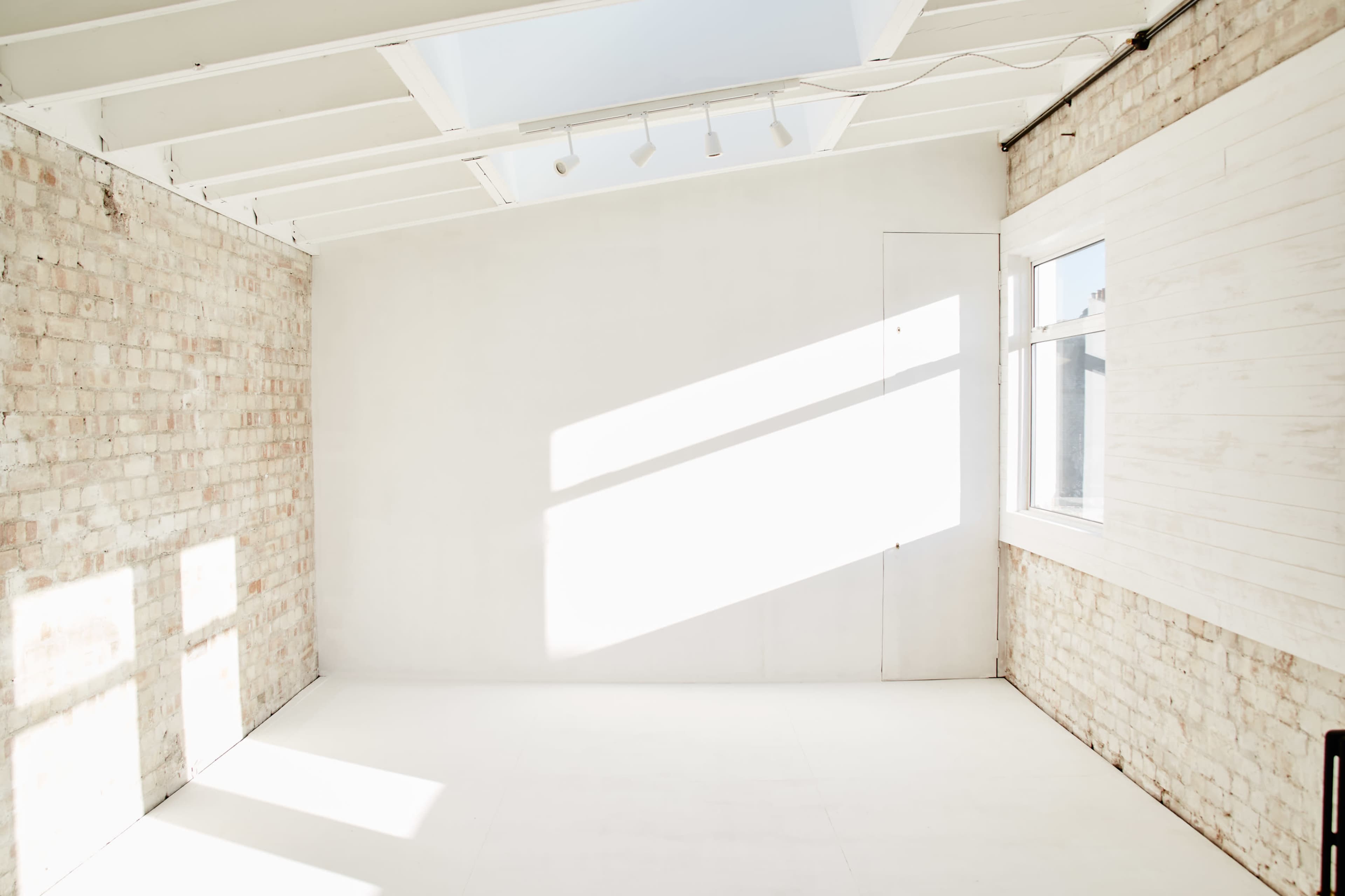 The image shows a bright, empty room with white walls and a partially brick wall, illuminated by natural light from a window.