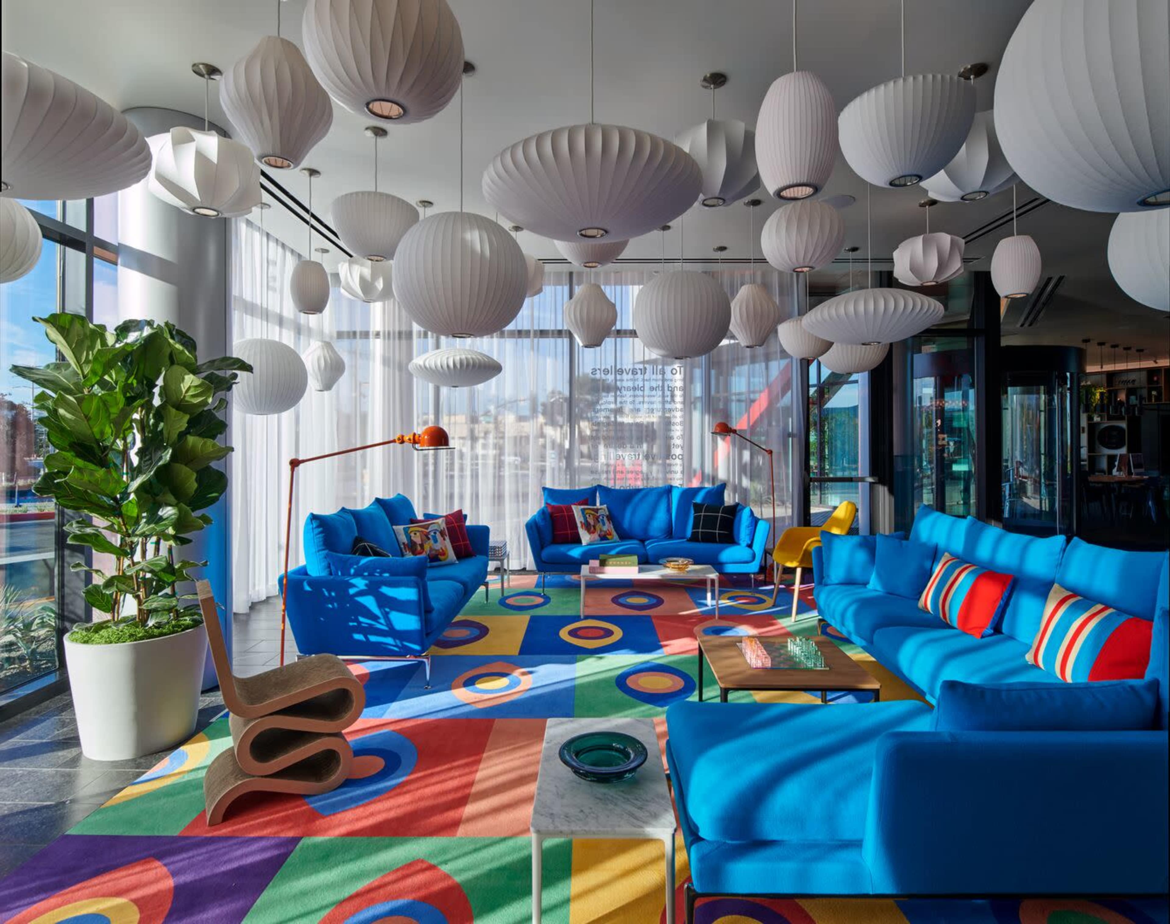 The image shows a modern lounge area featuring blue sofas, colorful circular-patterned carpets, and decorative paper lanterns hanging from the ceiling.