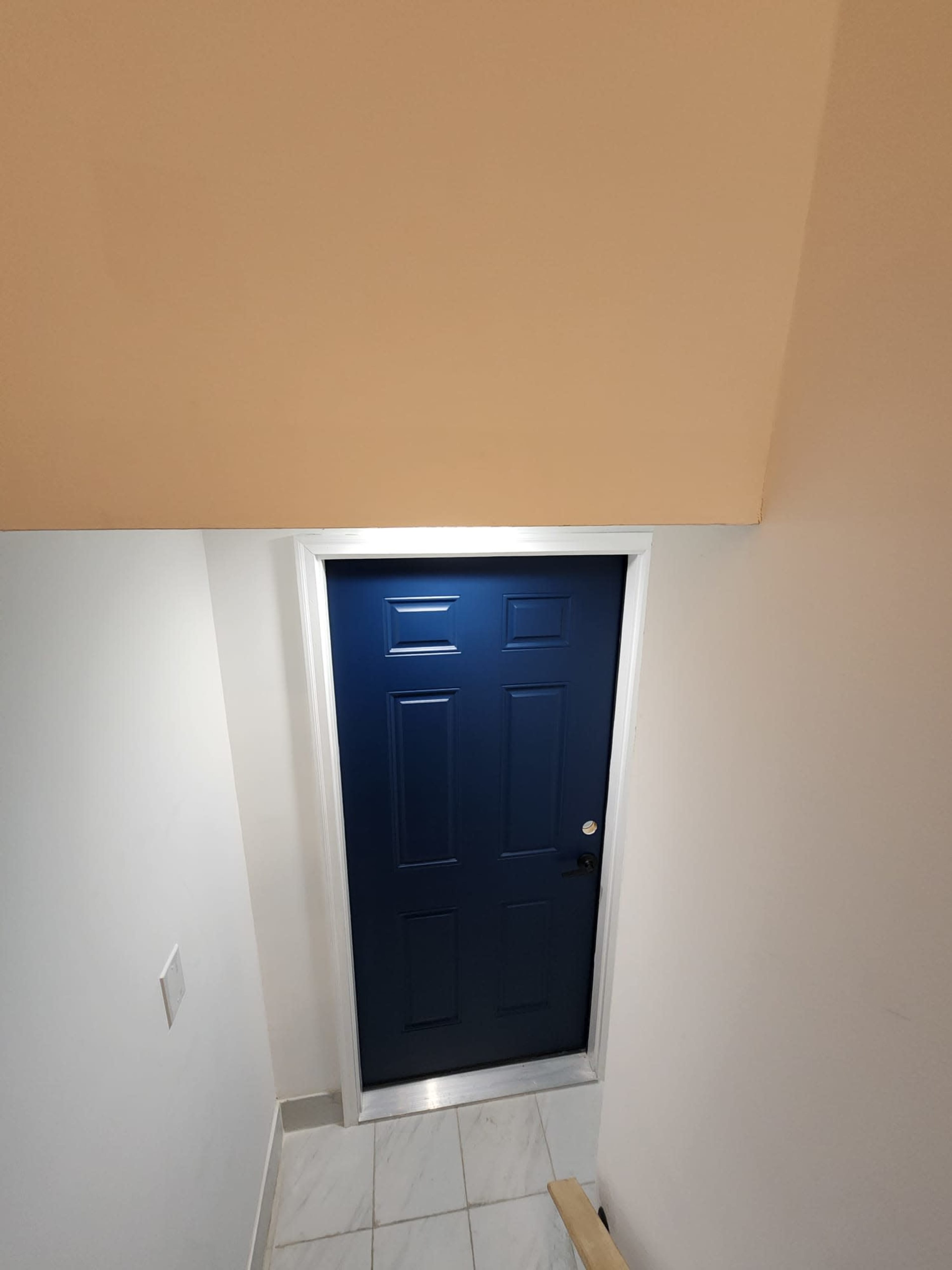 A blue door with six panels is located at the bottom of a staircase, framed by white walls and a ceiling.