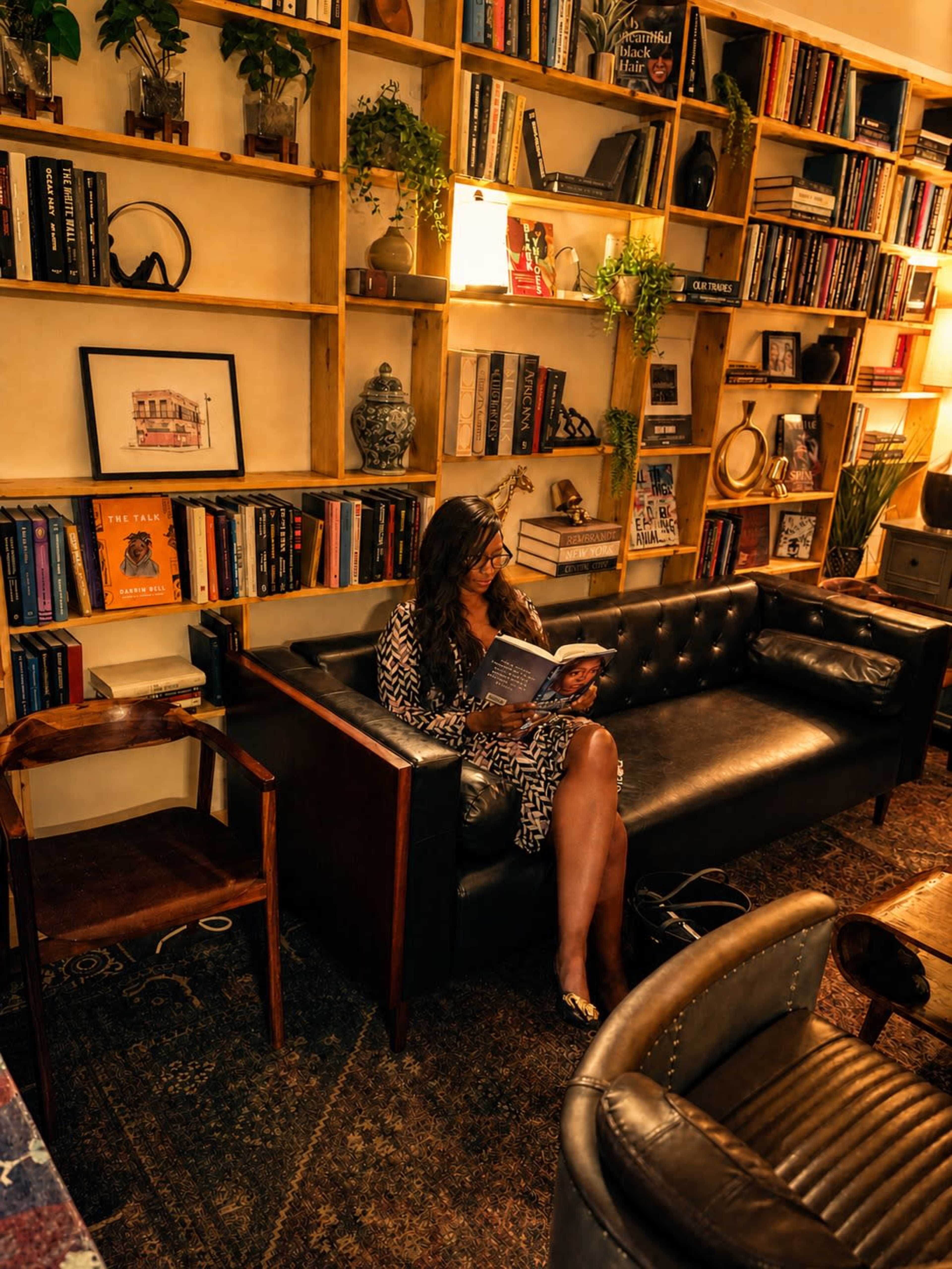 A woman sits on a black leather couch in a cozy library, reading a book surrounded by wooden shelves filled with books and plants.