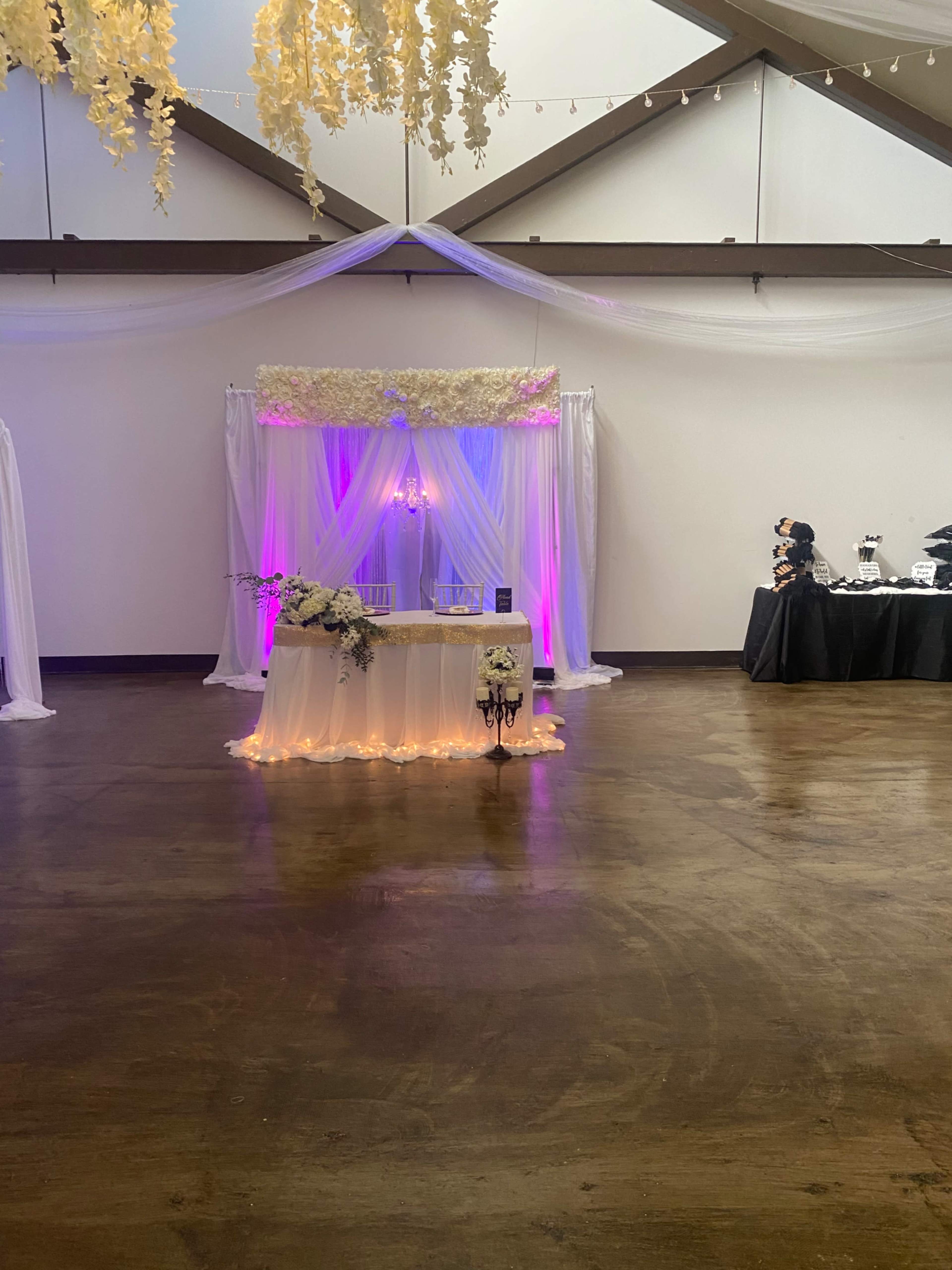 A decorated event space features a floral draped backdrop with soft lighting, a table at the center adorned with flowers, and additional setup in the background.