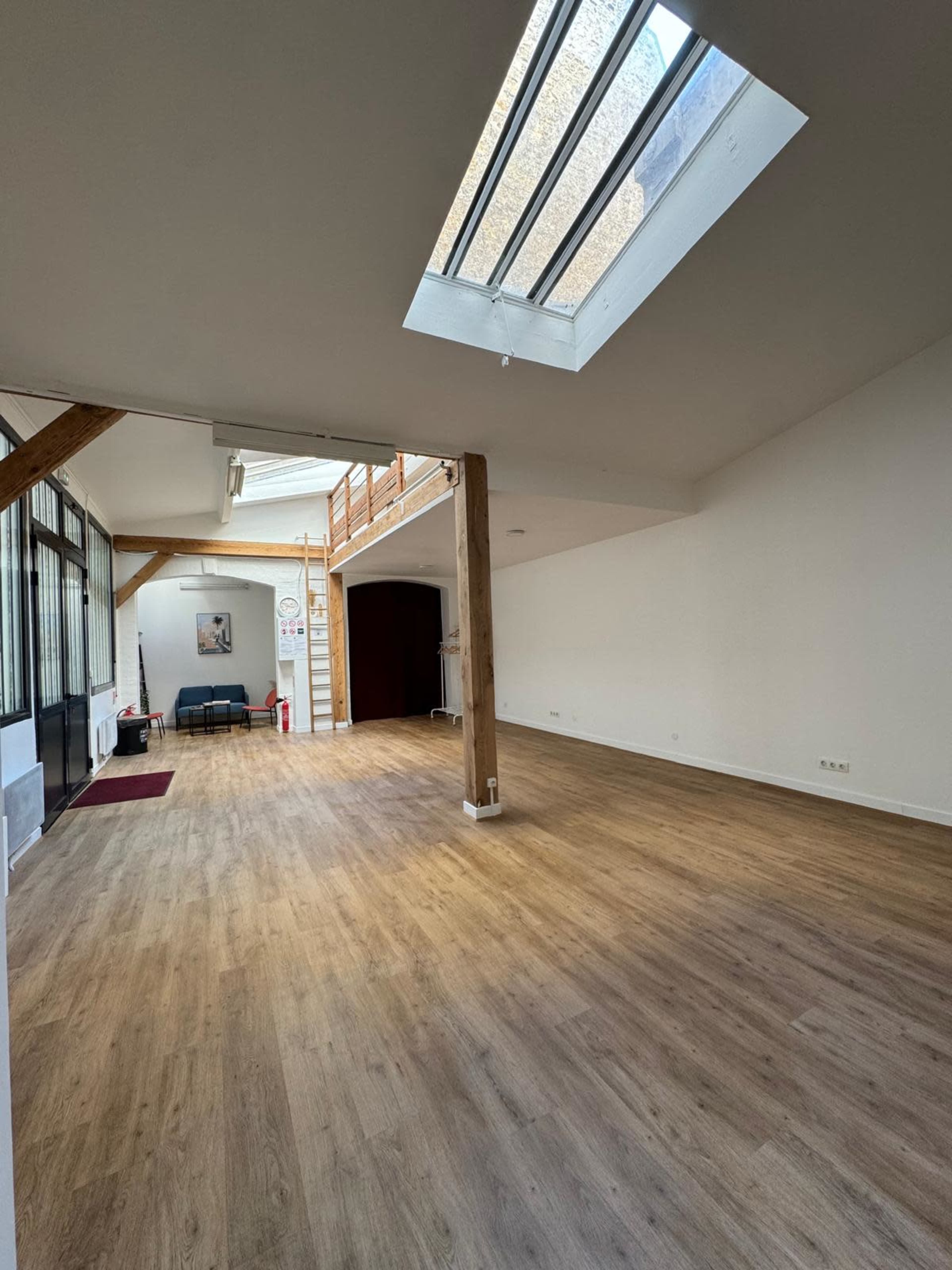 The image shows a spacious, empty interior with wooden flooring, large skylights, and wooden beams.