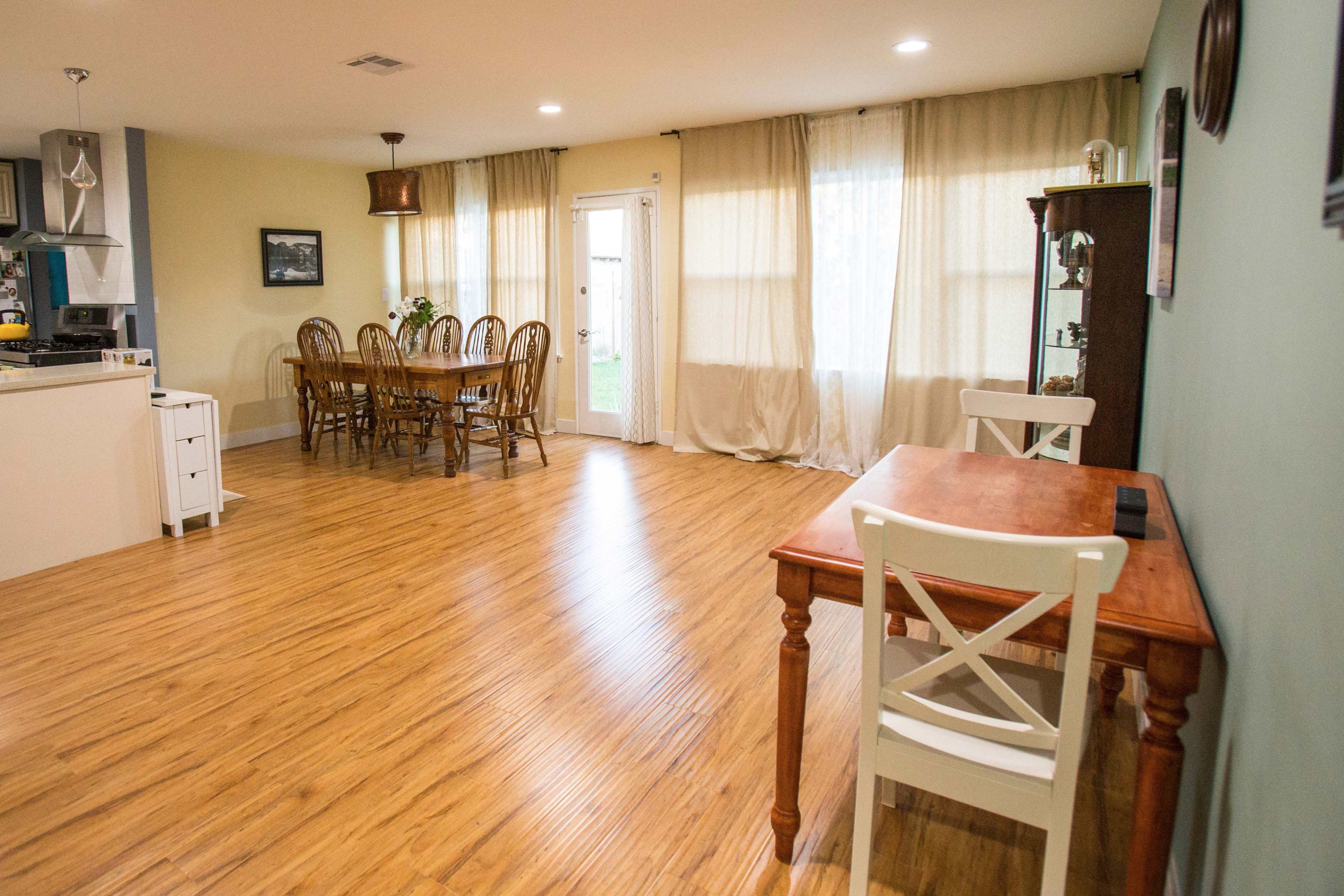 A spacious living area featuring a dining table set for six, a small desk near a window, and light-colored curtains.