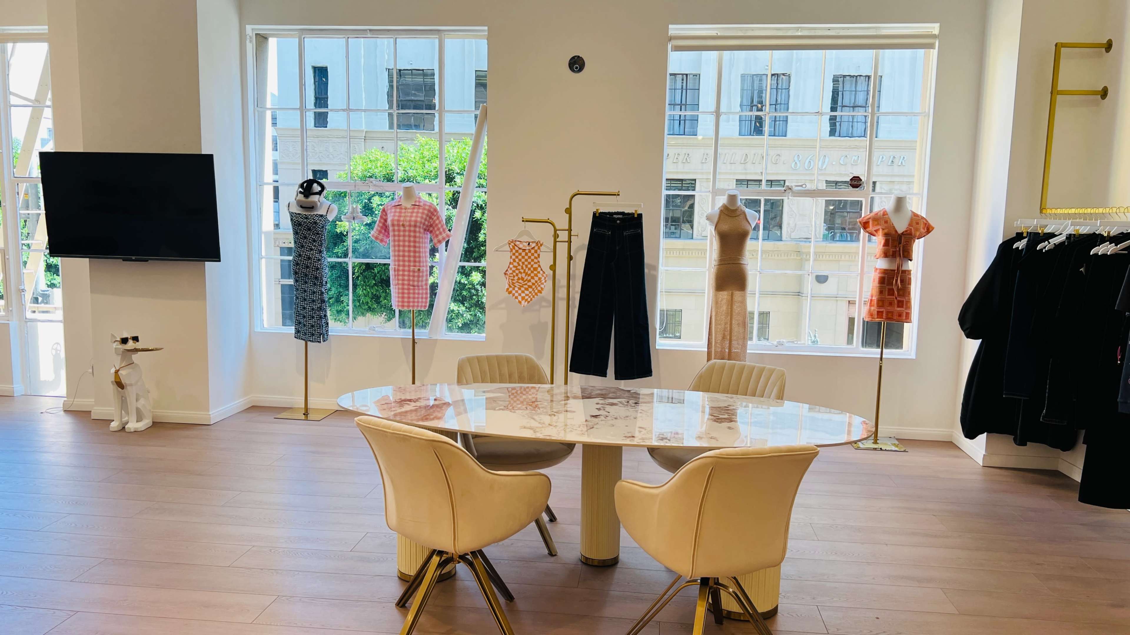 The image shows a well-lit boutique interior with a round table and upholstered chairs surrounded by mannequins displaying various clothing items.