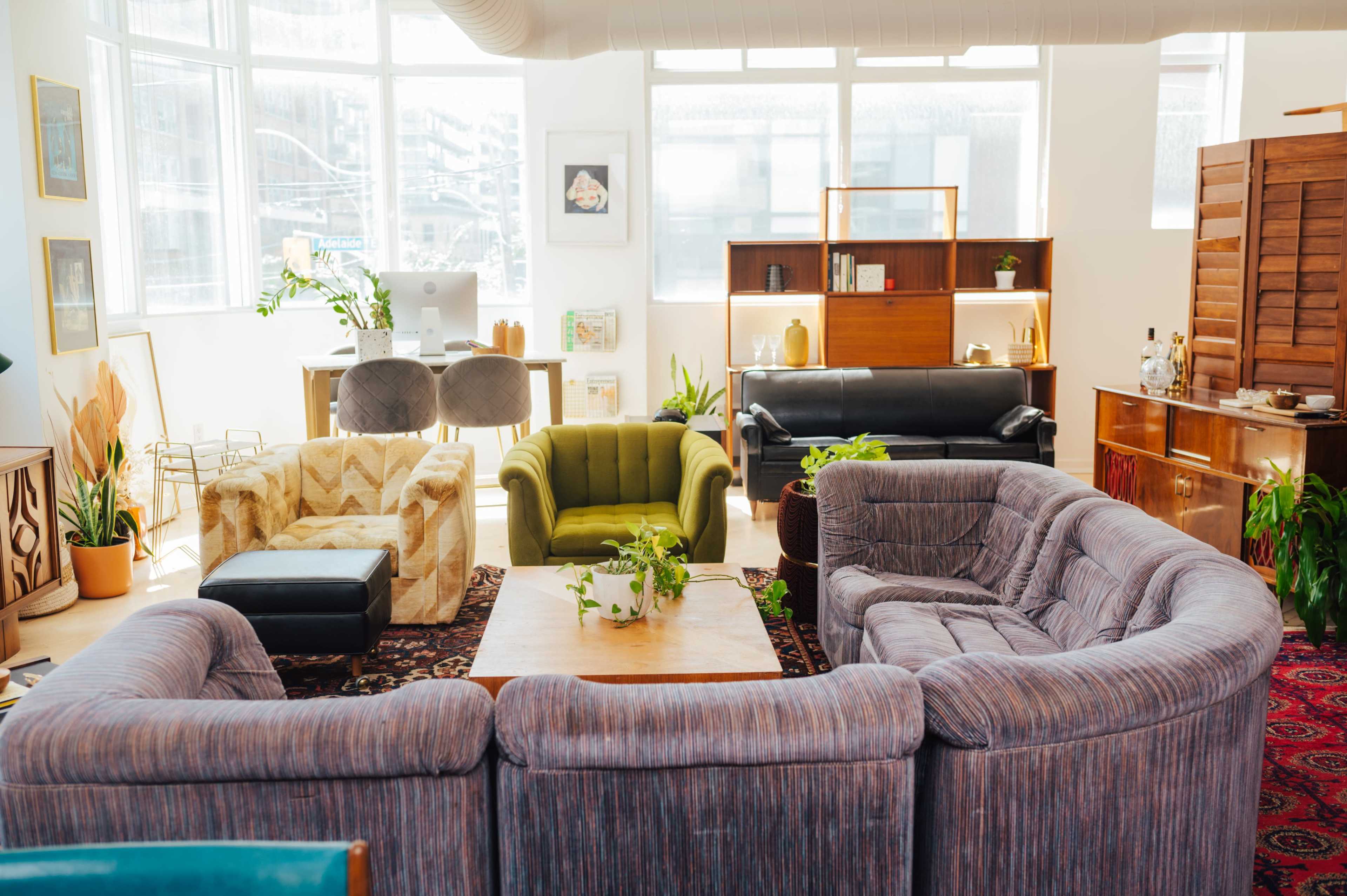The image shows a cozy, modern living space with a circular arrangement of sofas around a wooden table, complemented by various plants and a workspace in the background.