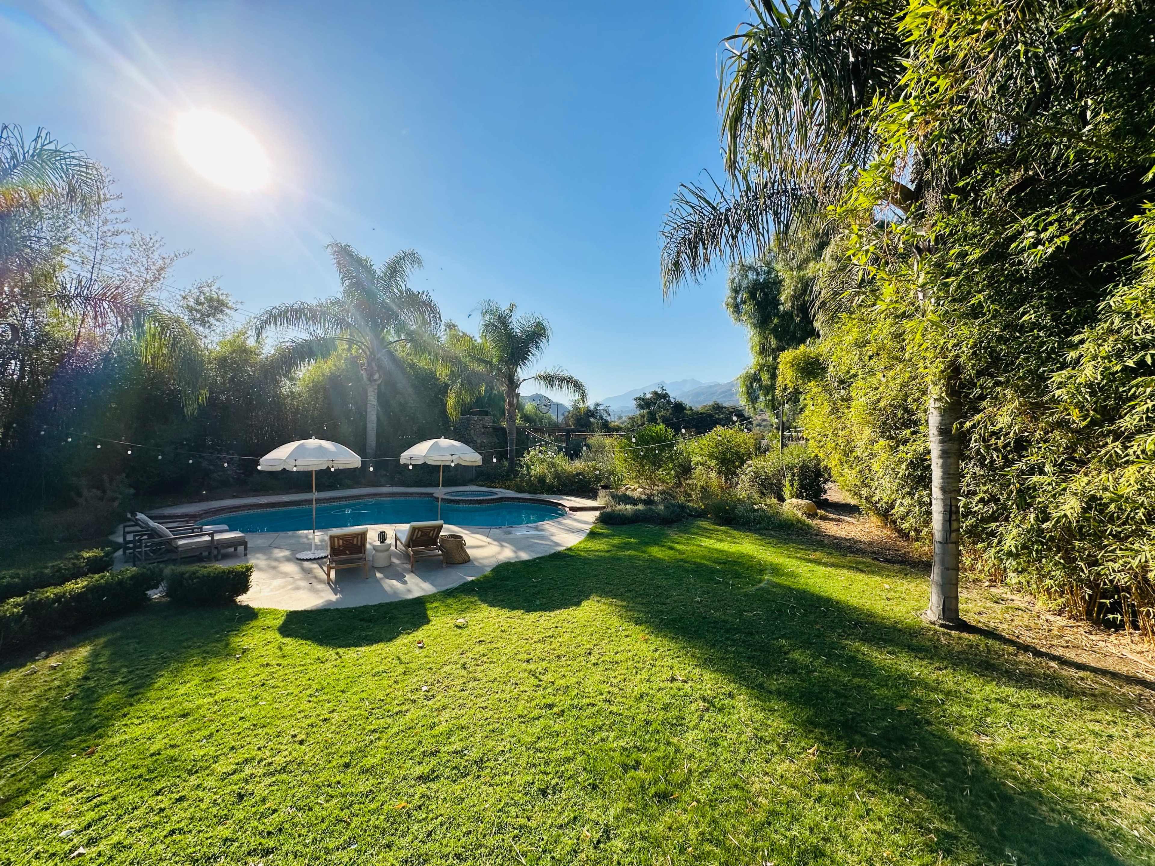 The image shows a backyard with a swimming pool, two lounge chairs under umbrellas, and surrounding lush greenery.
