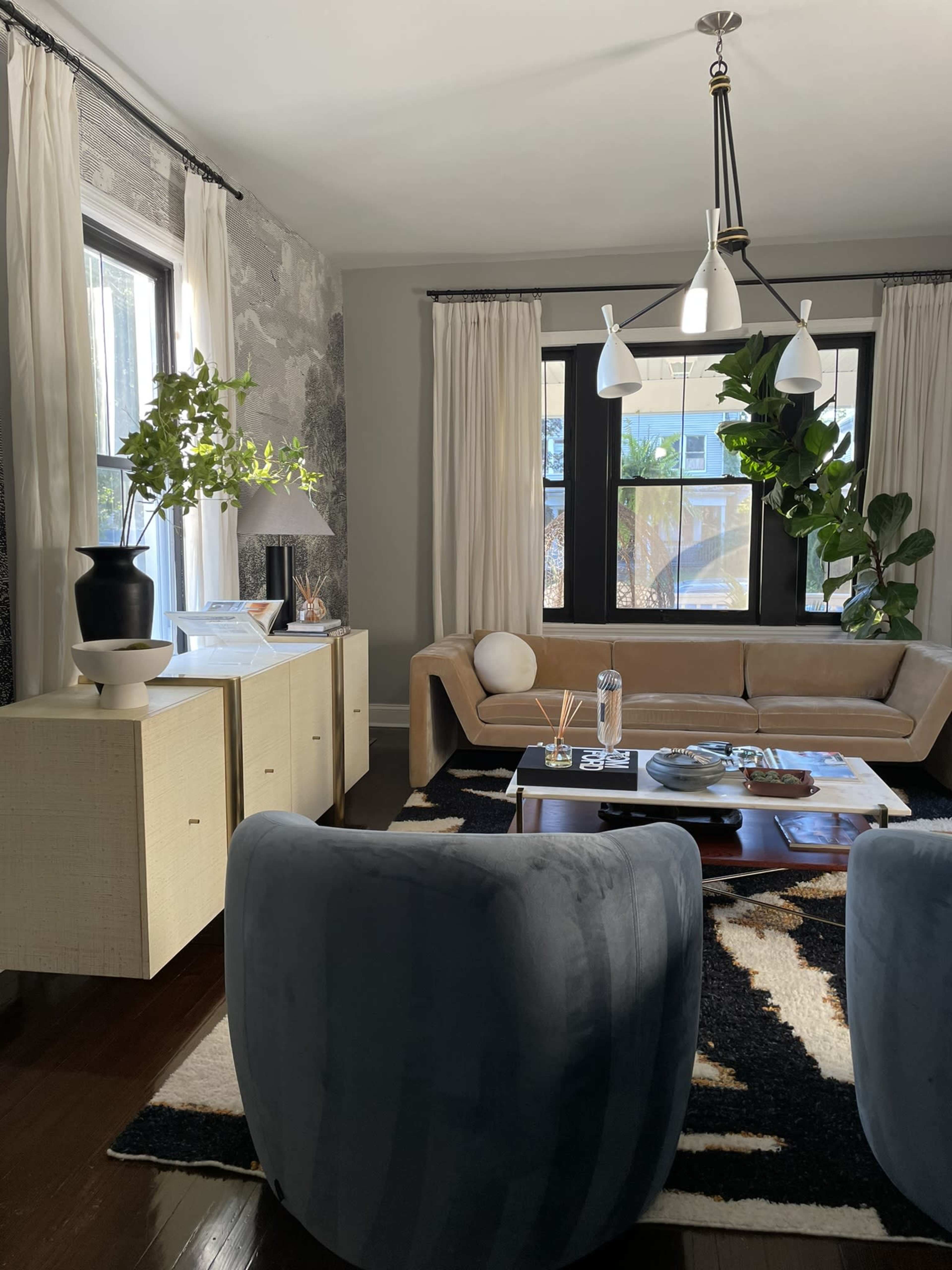 A living room features a beige sofa, round blue chairs, a coffee table with magazines, and a stylish decor of plants and lighting.