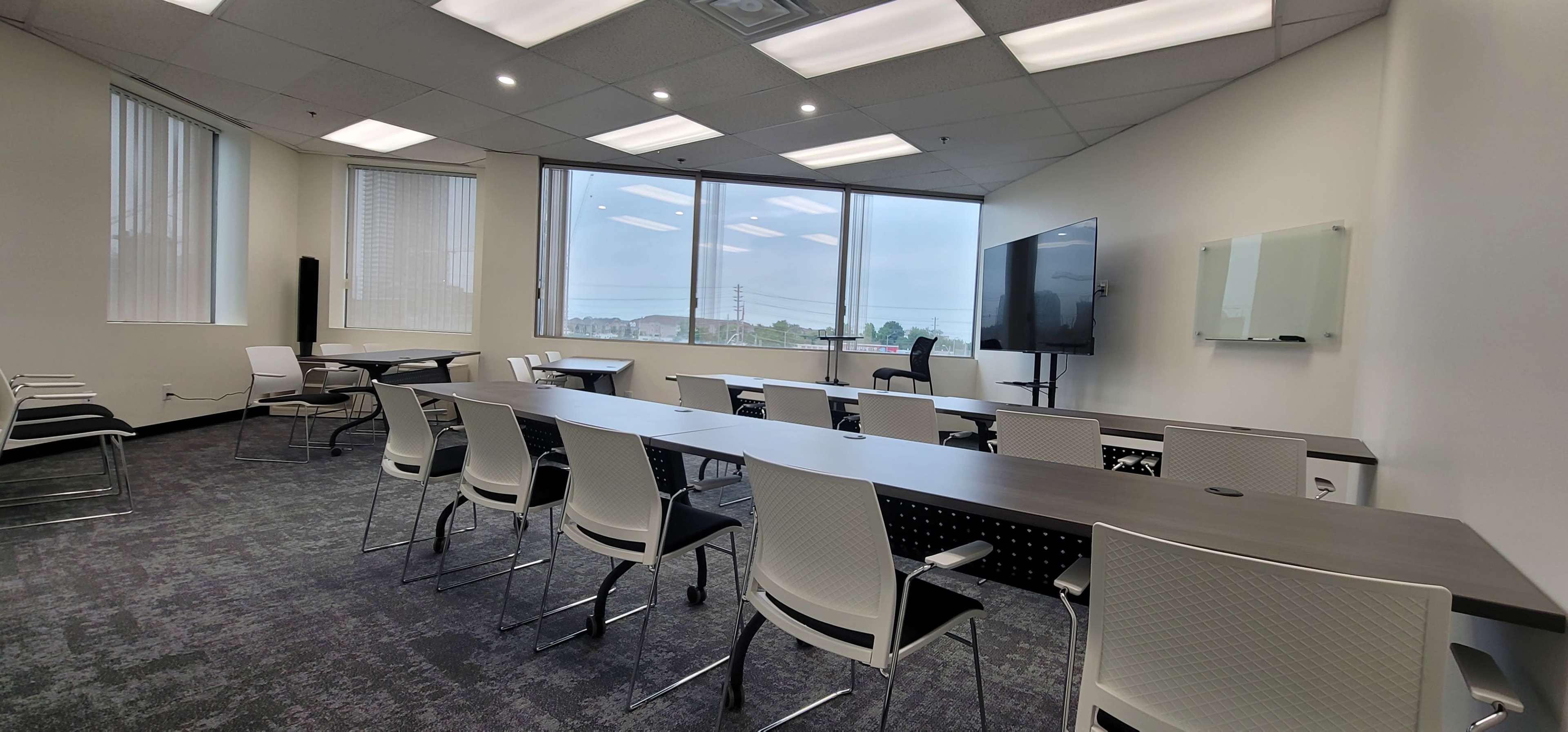 A modern conference room equipped with a long table, chairs, a large screen, and windows offering a view of the outdoors.