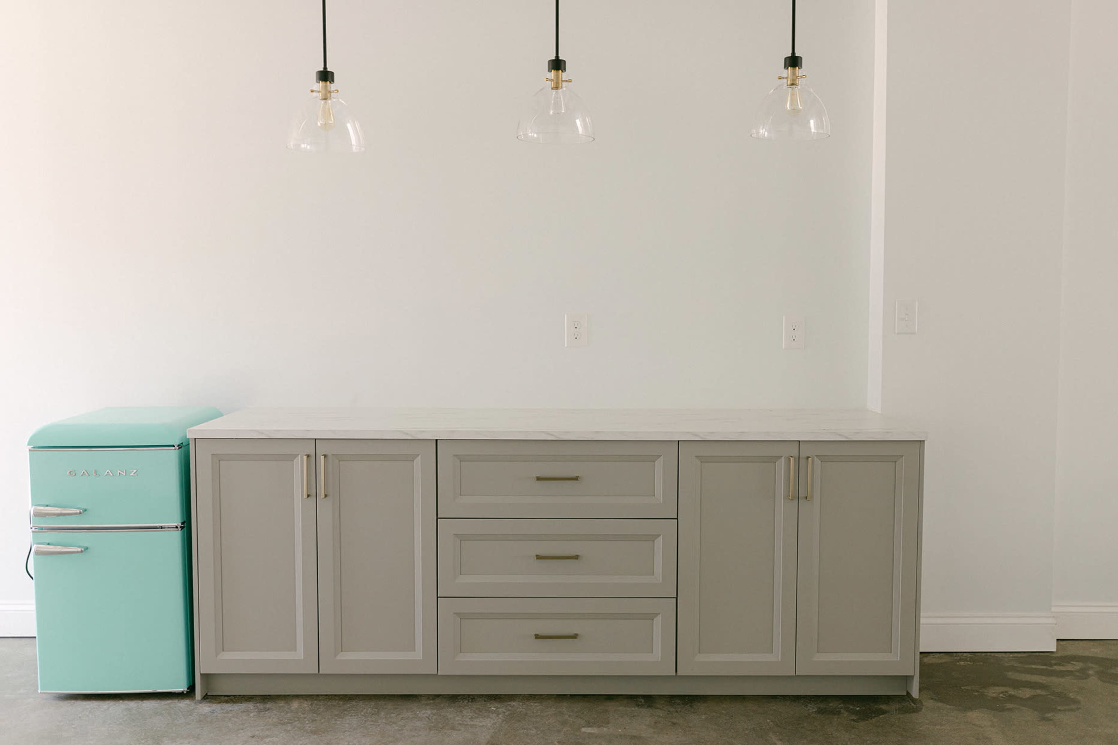 A kitchen setup features a light gray cabinet with three drawers and a matching countertop next to a mint green retro-style refrigerator, illuminated by three pendant lights.