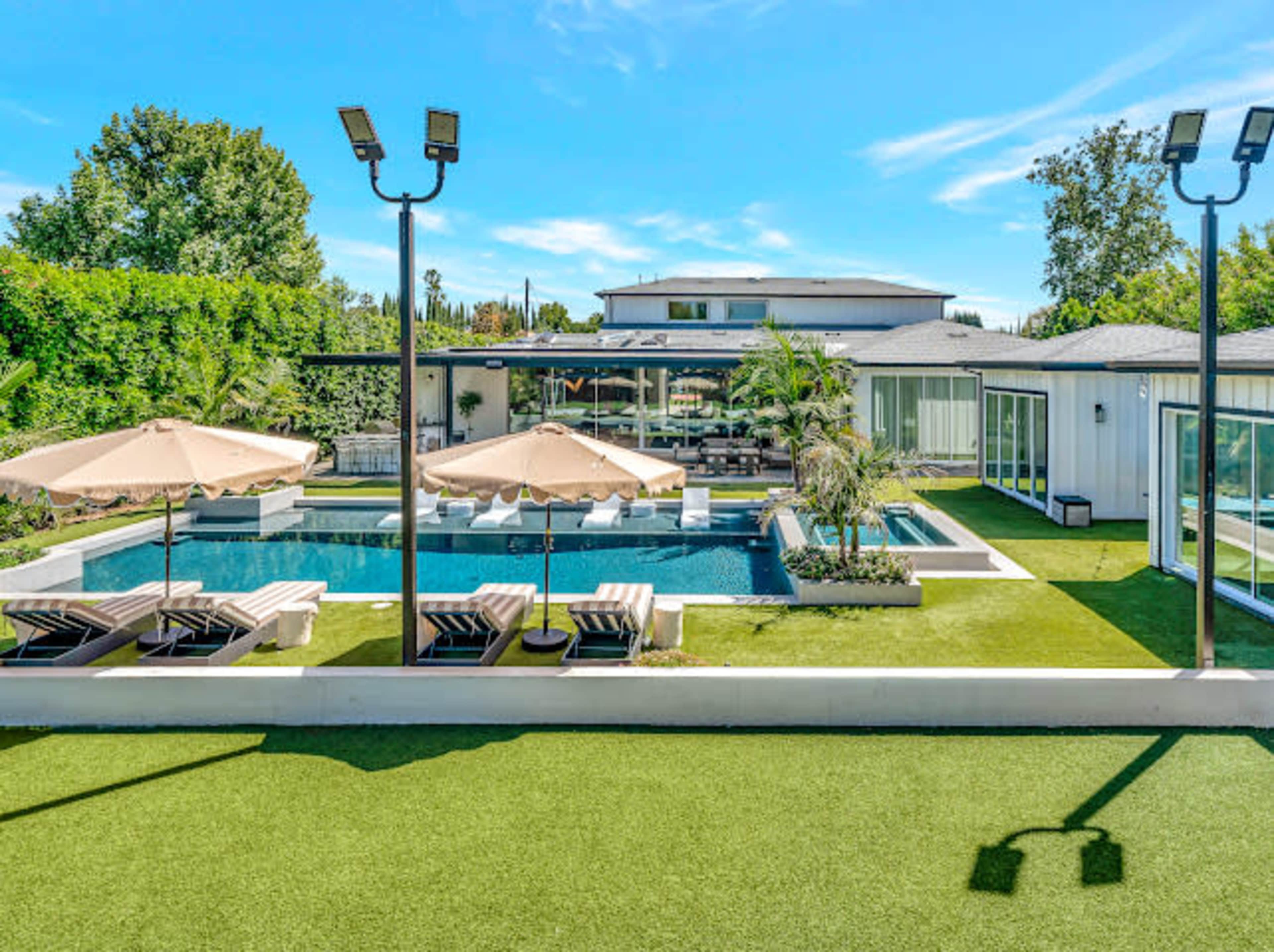The image shows a modern home with a spacious outdoor area featuring a swimming pool, lounge chairs, and umbrellas, surrounded by greenery and well-maintained landscaping.