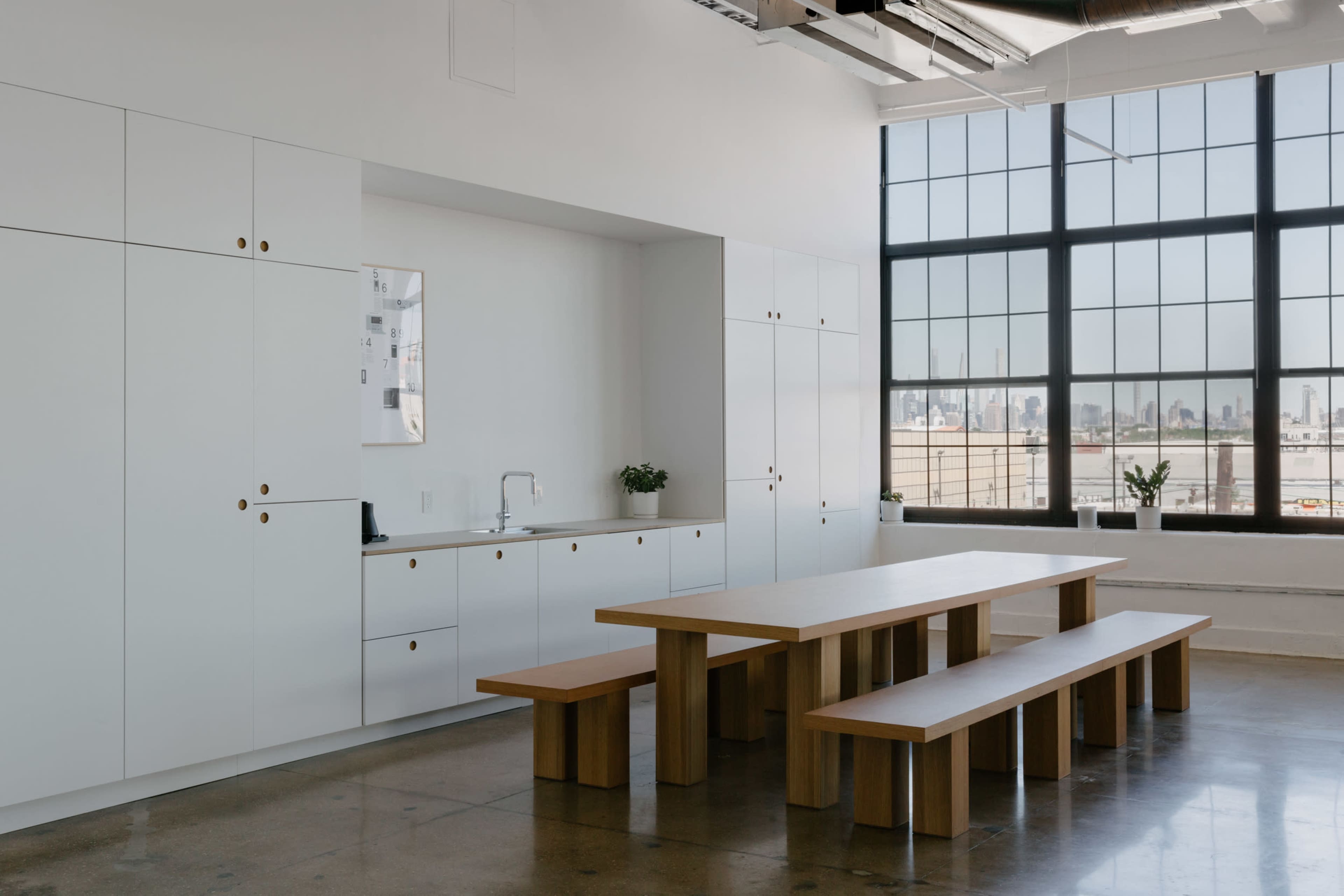 A modern kitchen area features white cabinetry, a large wooden table with benches, and a wide window overlooking a city skyline.