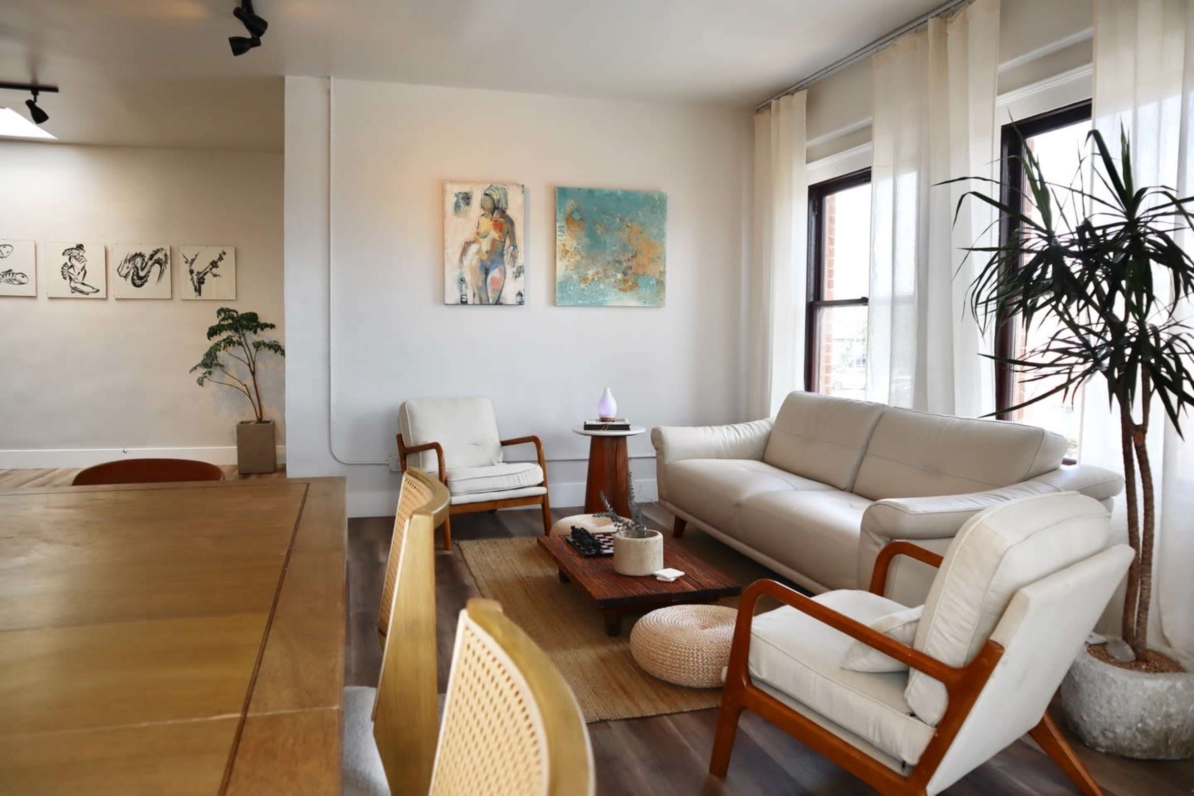 The image shows a modern living room with a white sofa, wooden chairs, a coffee table, and large windows draped with light curtains.