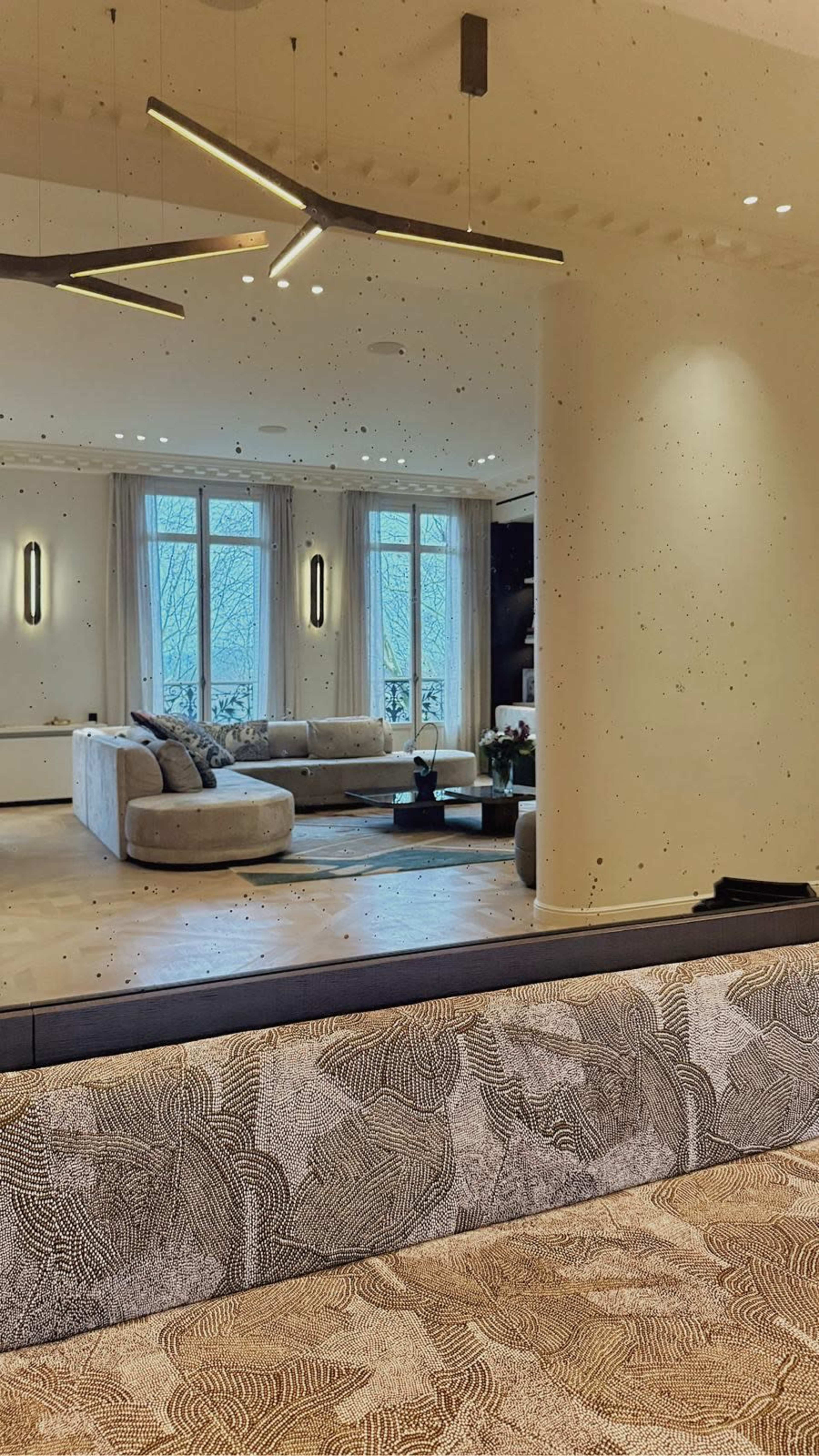 A spacious living room is reflected in a large mirror, featuring a light-colored sectional sofa, a coffee table, and large windows allowing natural light to enter.