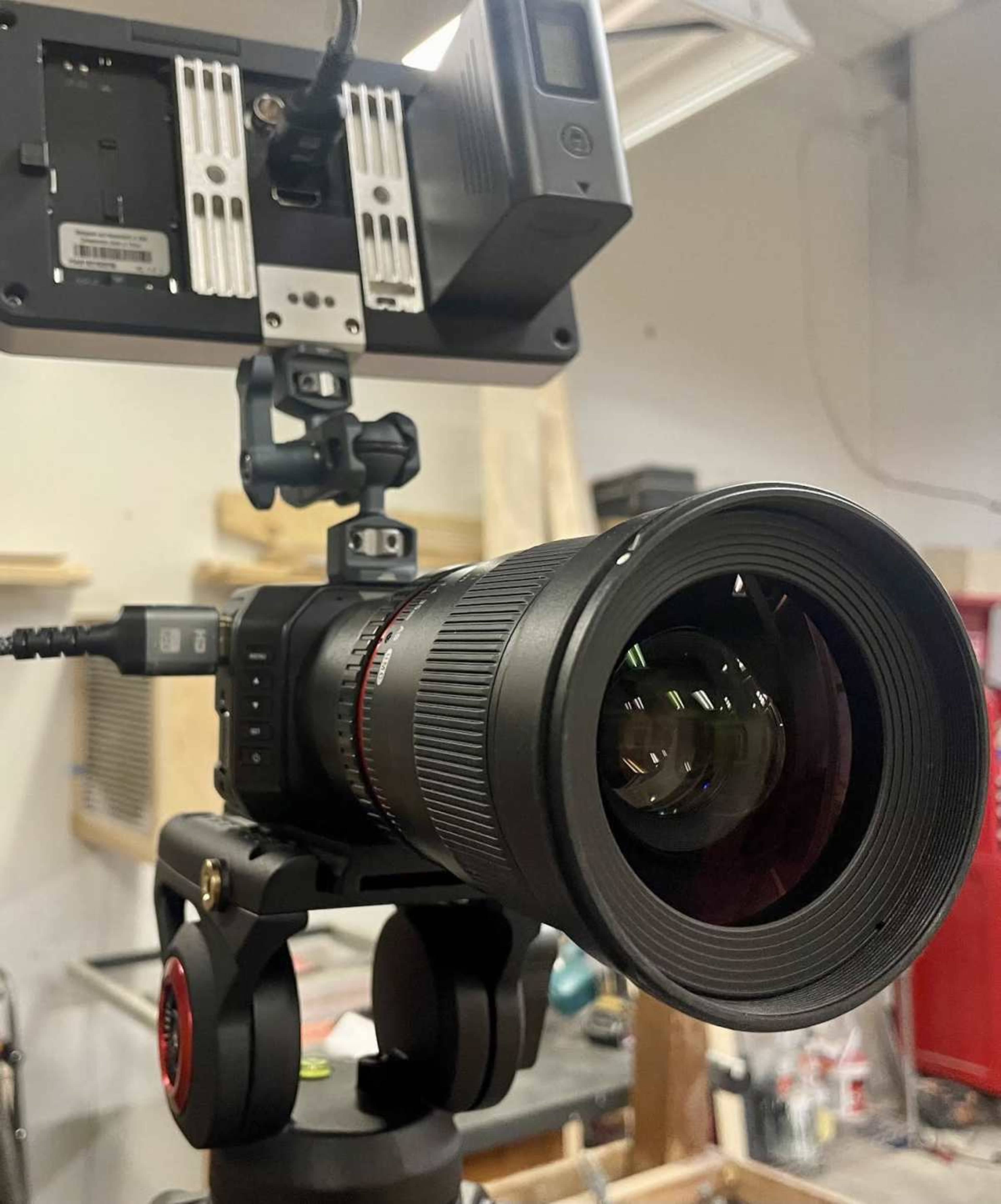 A camera with a large lens is mounted on a tripod, with an attached monitor and additional accessories in a workshop setting.