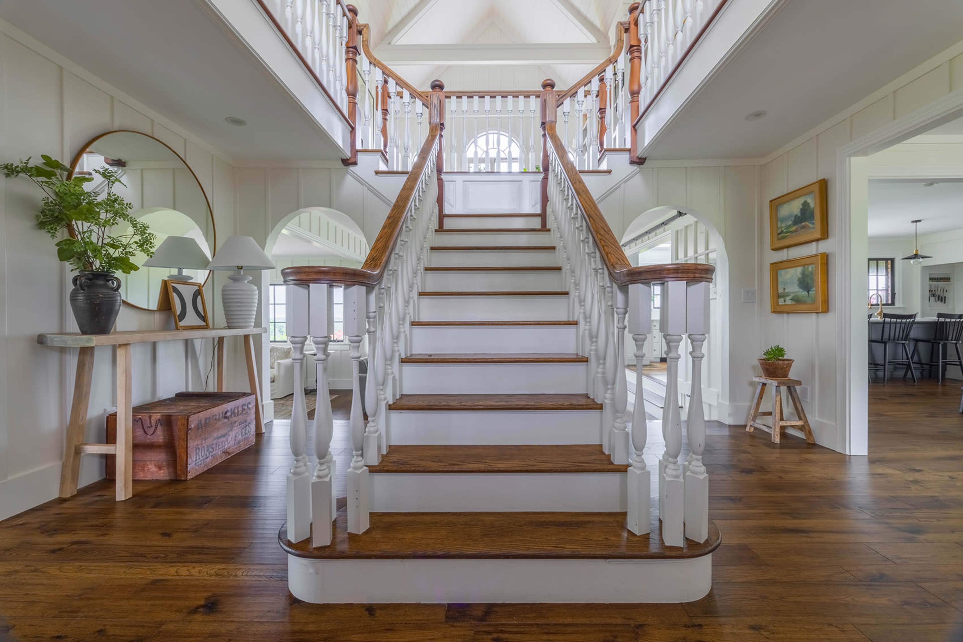 The image shows a bright interior with a wooden staircase leading to an upper level, surrounded by white walls and elegant railings.