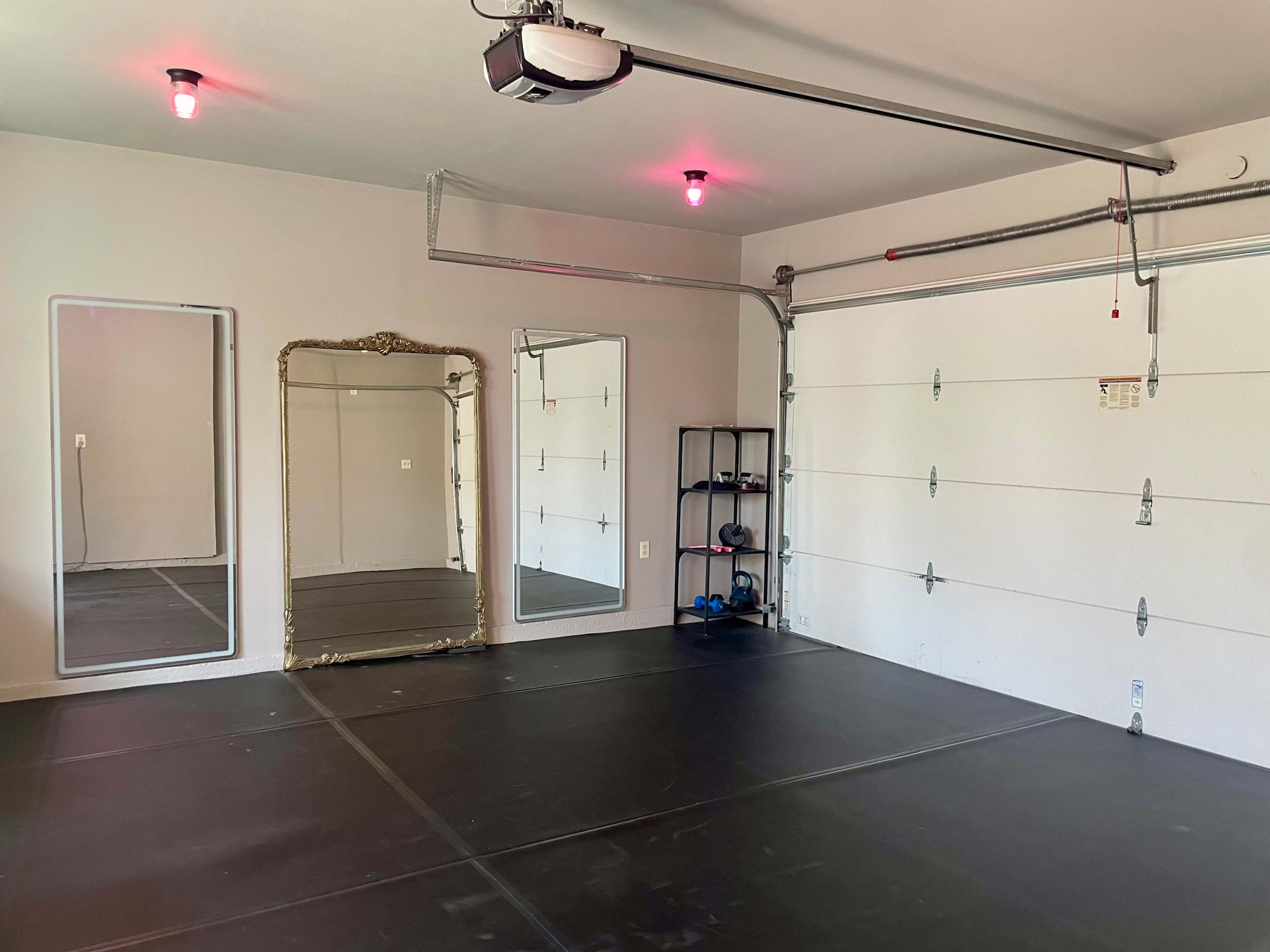 The image shows a spacious garage with a smooth black floor, a large mirror, and a shelf holding exercise equipment.
