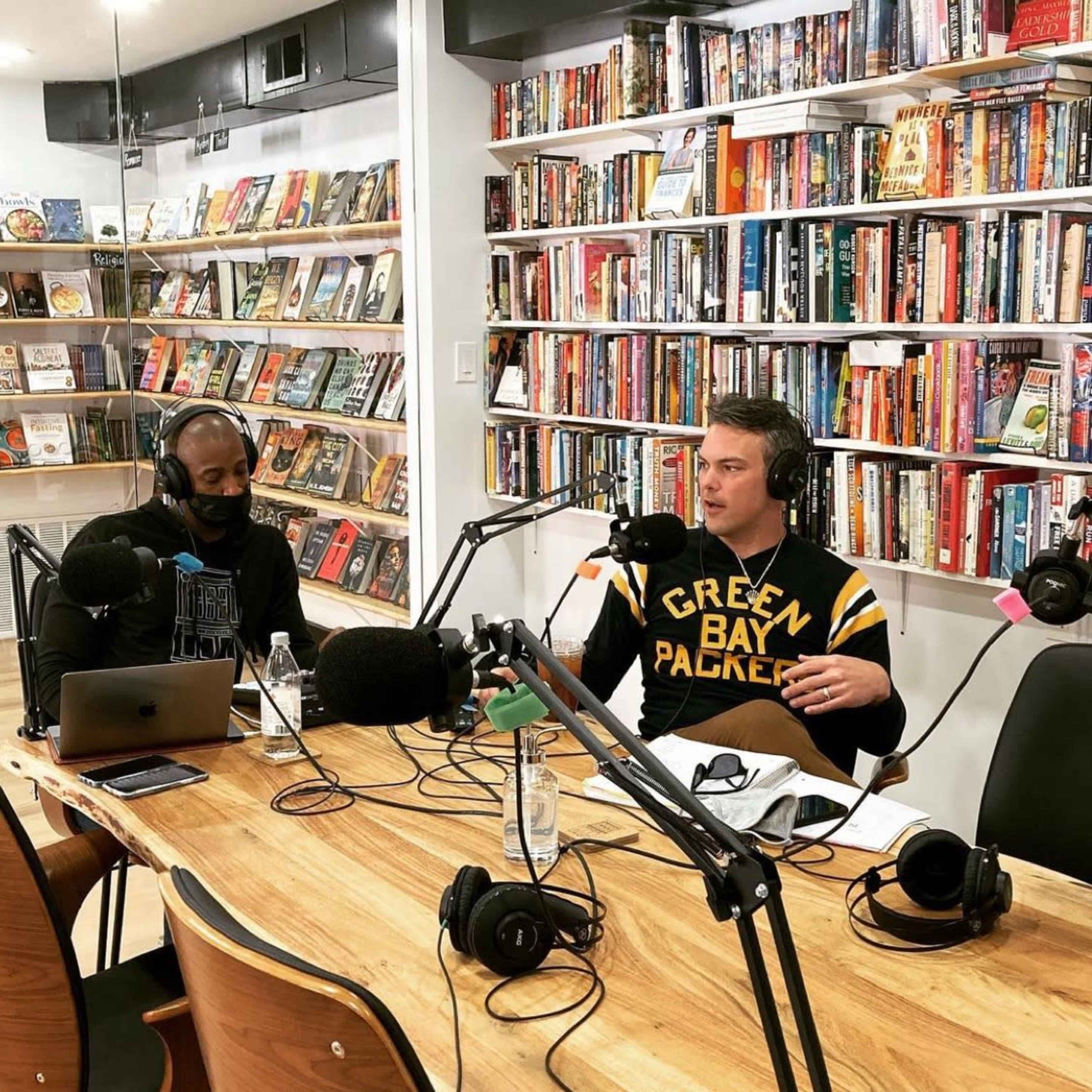 Two men are seated at a wooden table with microphones and laptops in front of them, discussing in a brightly lit room lined with bookshelves filled with various books.