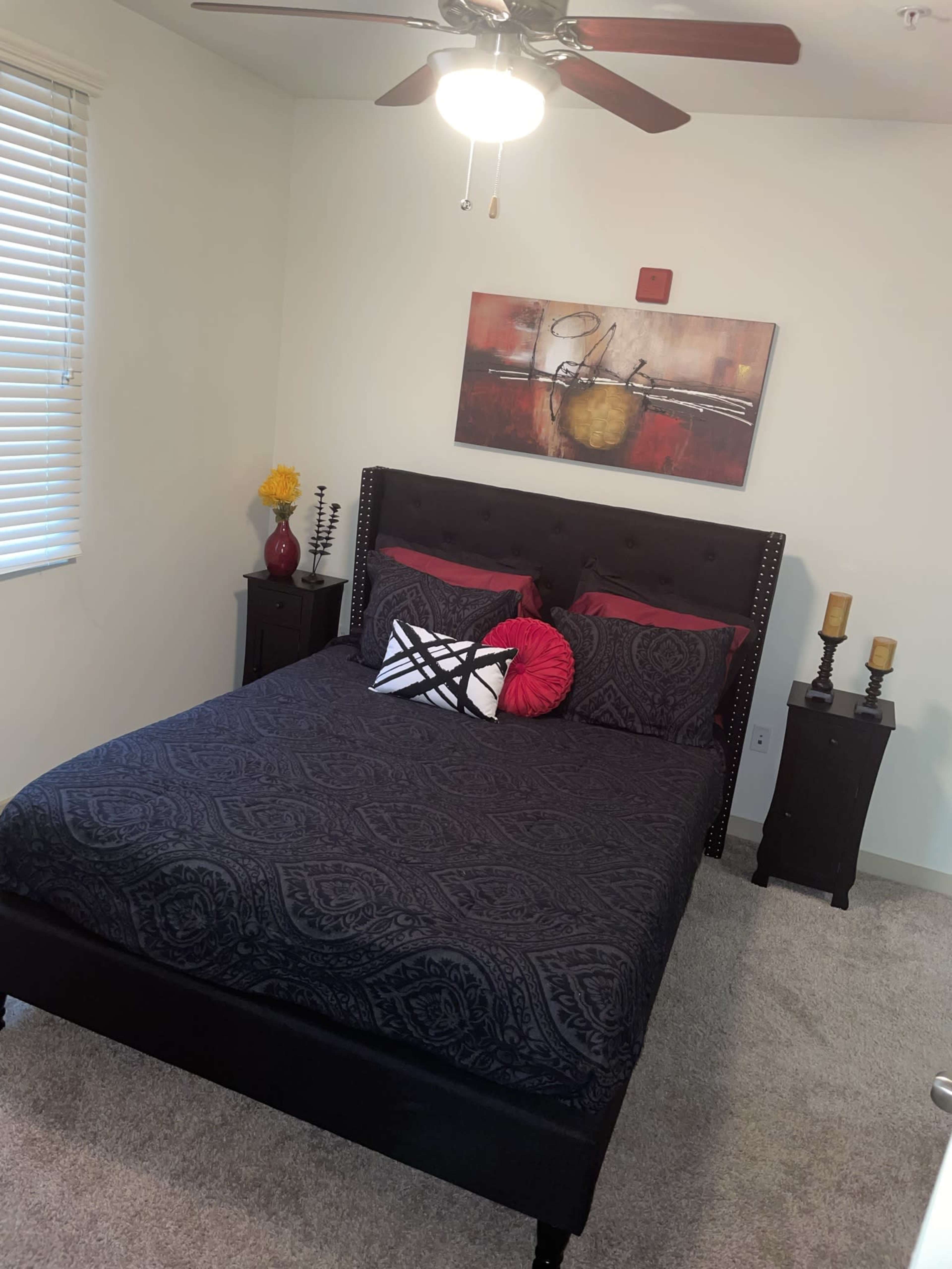 A neatly arranged bedroom features a black bedspread with decorative pillows, a ceiling fan, and decorative art on the wall, complemented by bedside tables.