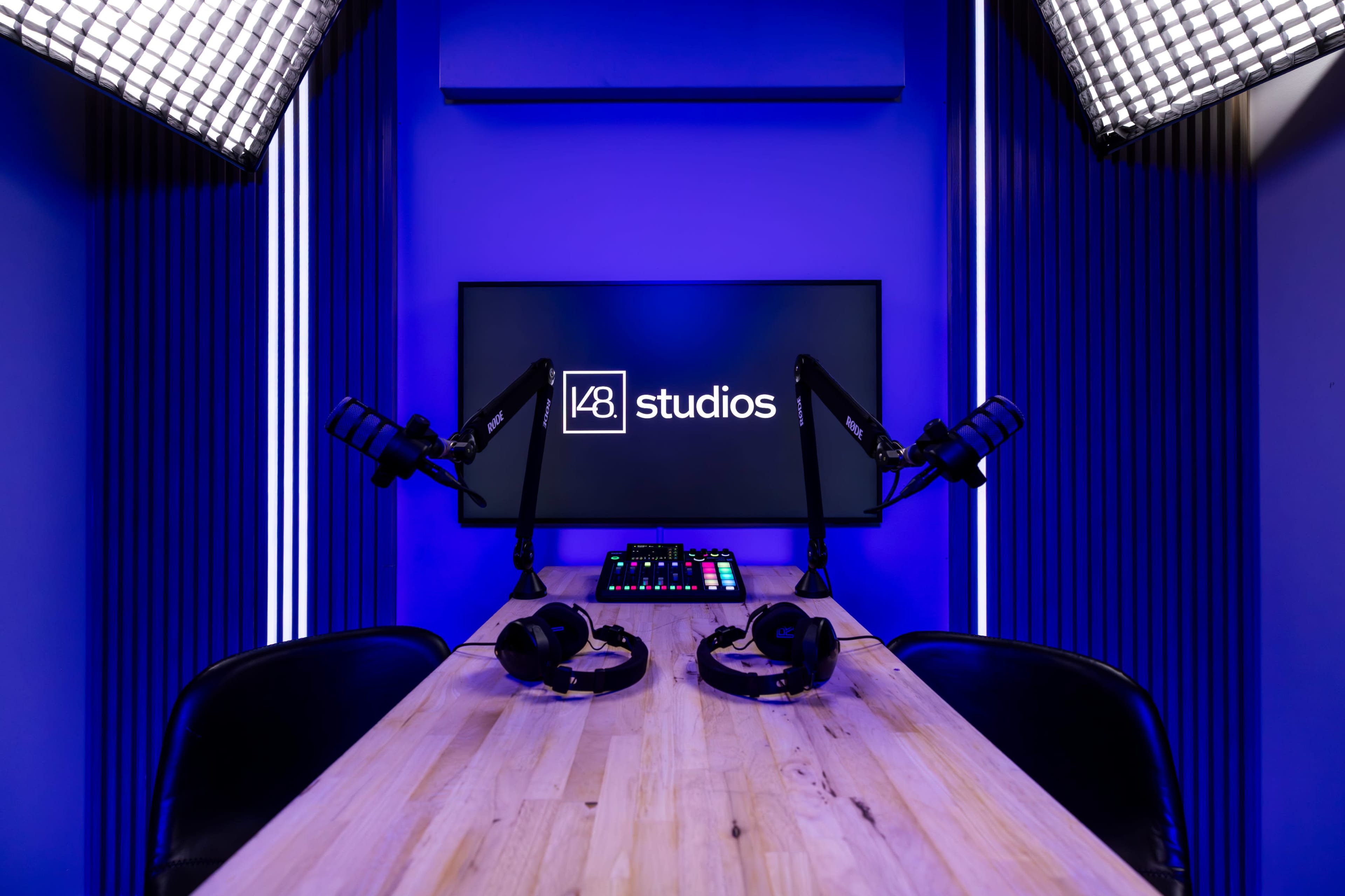 The image shows a modern podcast studio featuring a wooden table, two microphones on adjustable arms, headphones, a control panel, and a large screen that displays the logo "studios" against a purple-lit background.