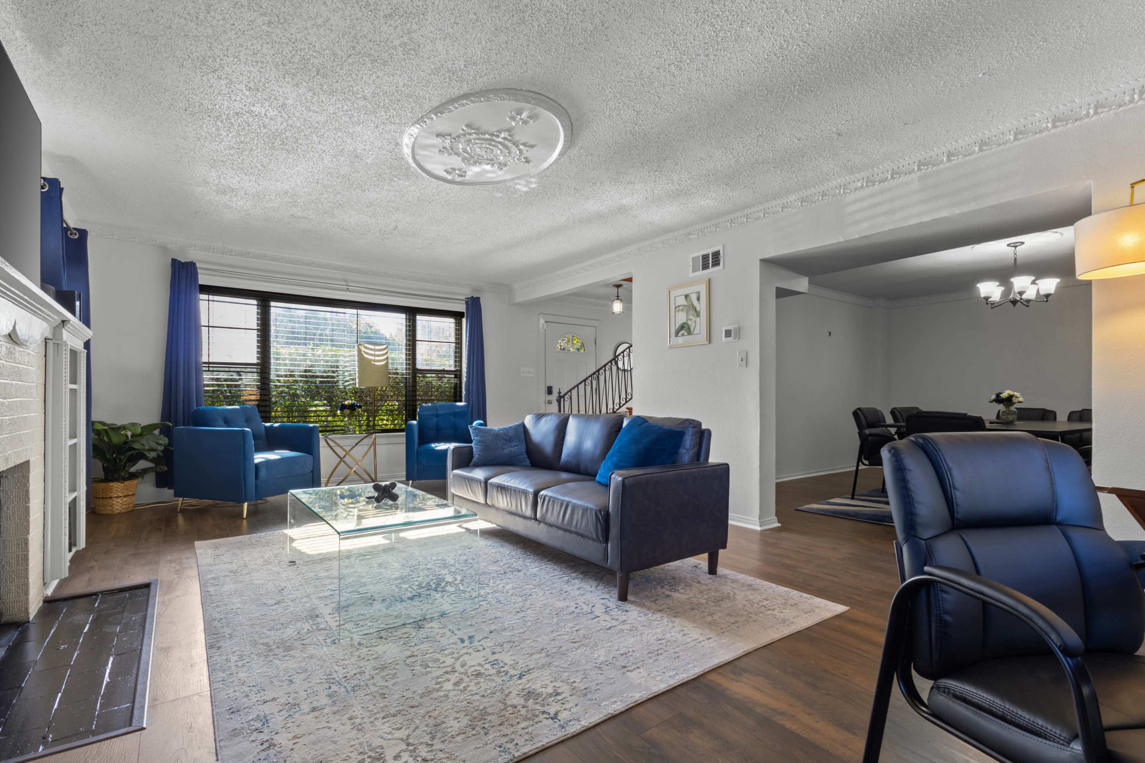 The living room features a sofa and armchairs in blue upholstery, a glass coffee table, and a view through large windows with curtains.
