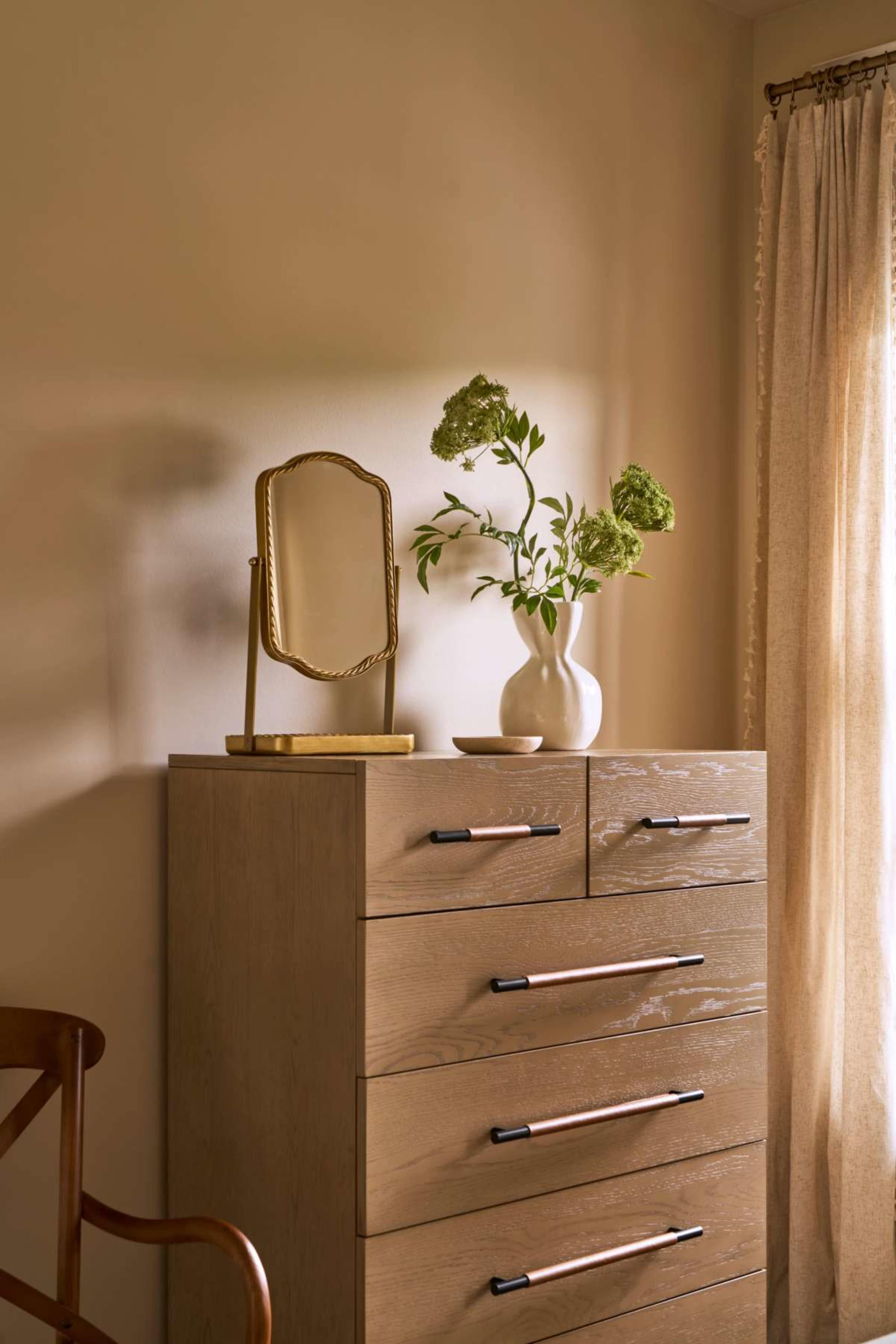 A wooden dresser with five drawers displays a small mirror and a vase of green flowers against a softly lit wall and a curtain.
