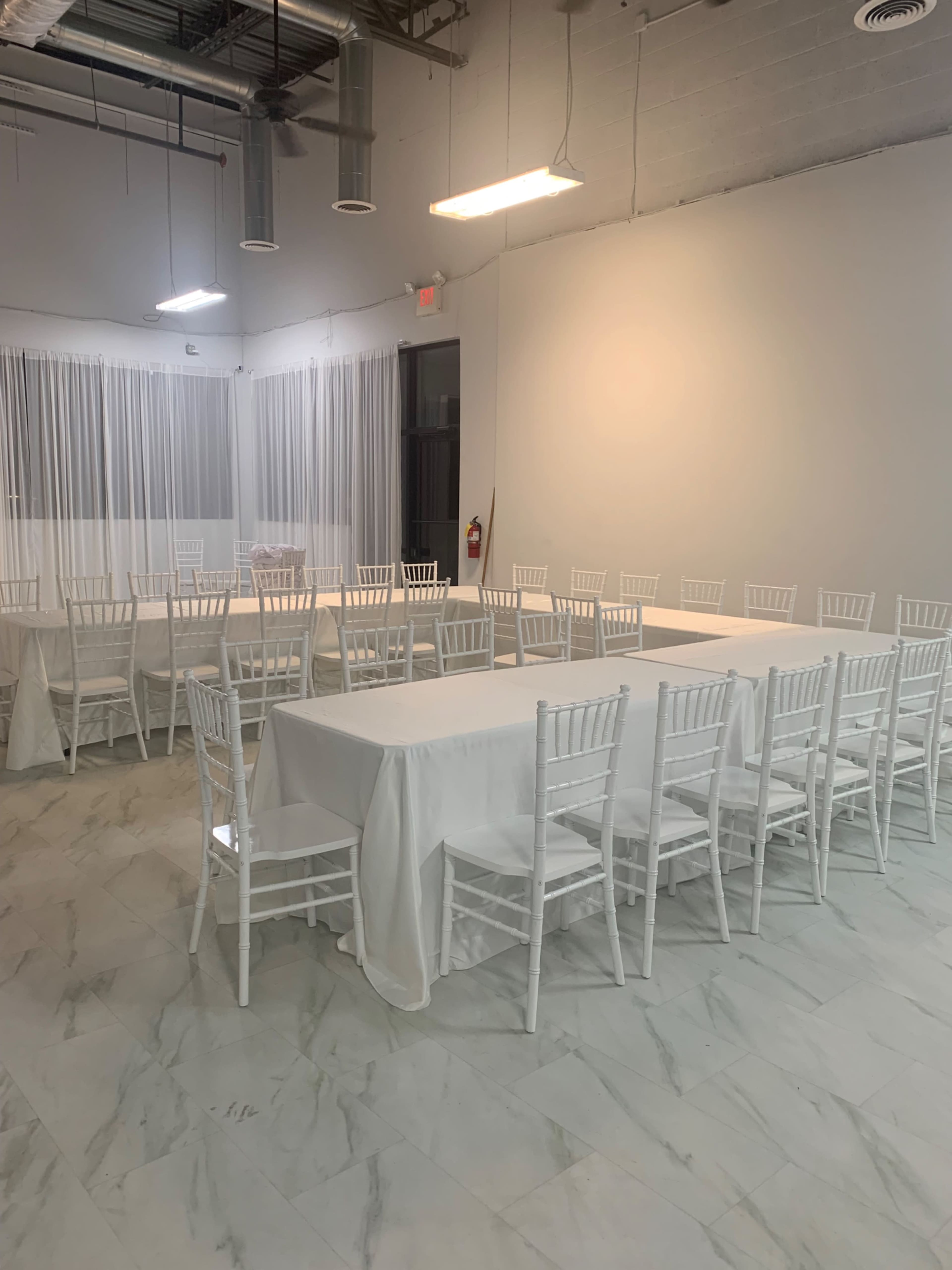 A spacious room with white walls and marble flooring, set up with multiple tables covered in white linens and surrounded by white chiavari chairs.