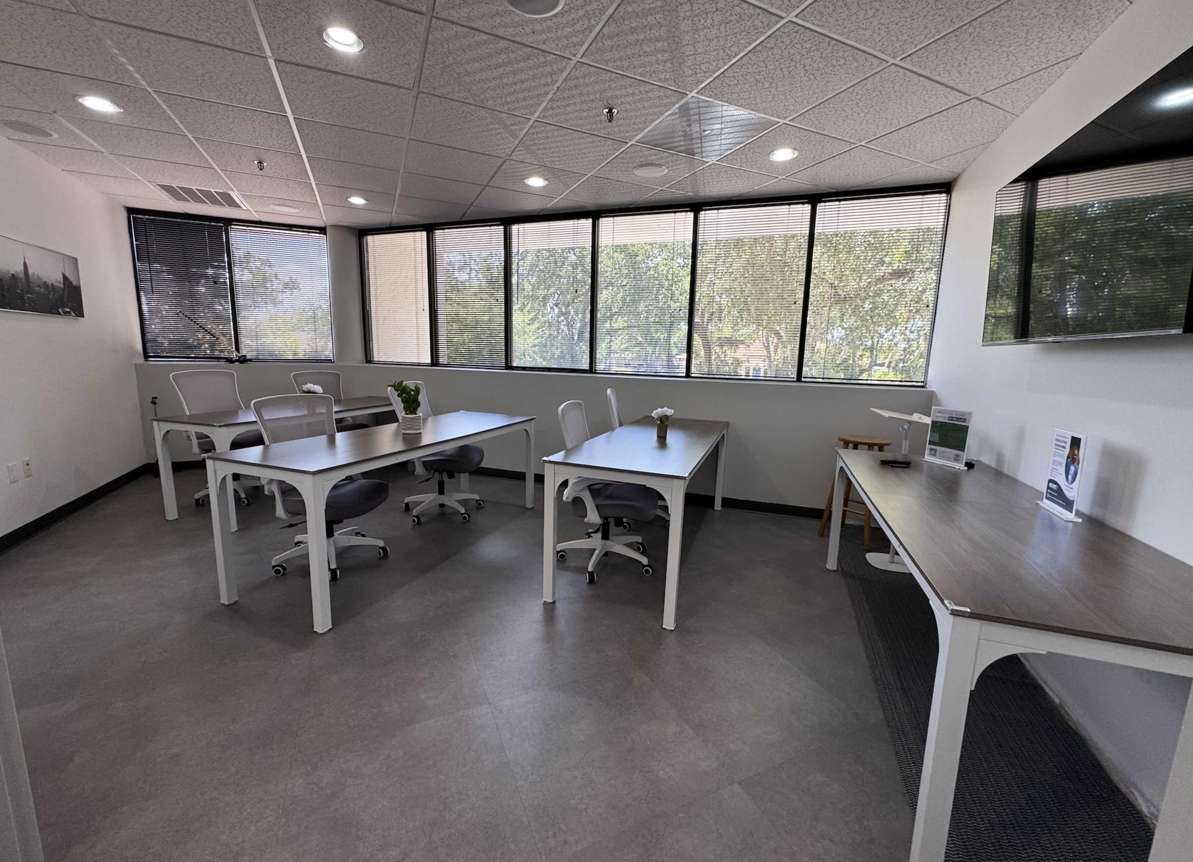 The image shows a bright, modern office space with several work desks, chairs, and large windows allowing natural light to enter.