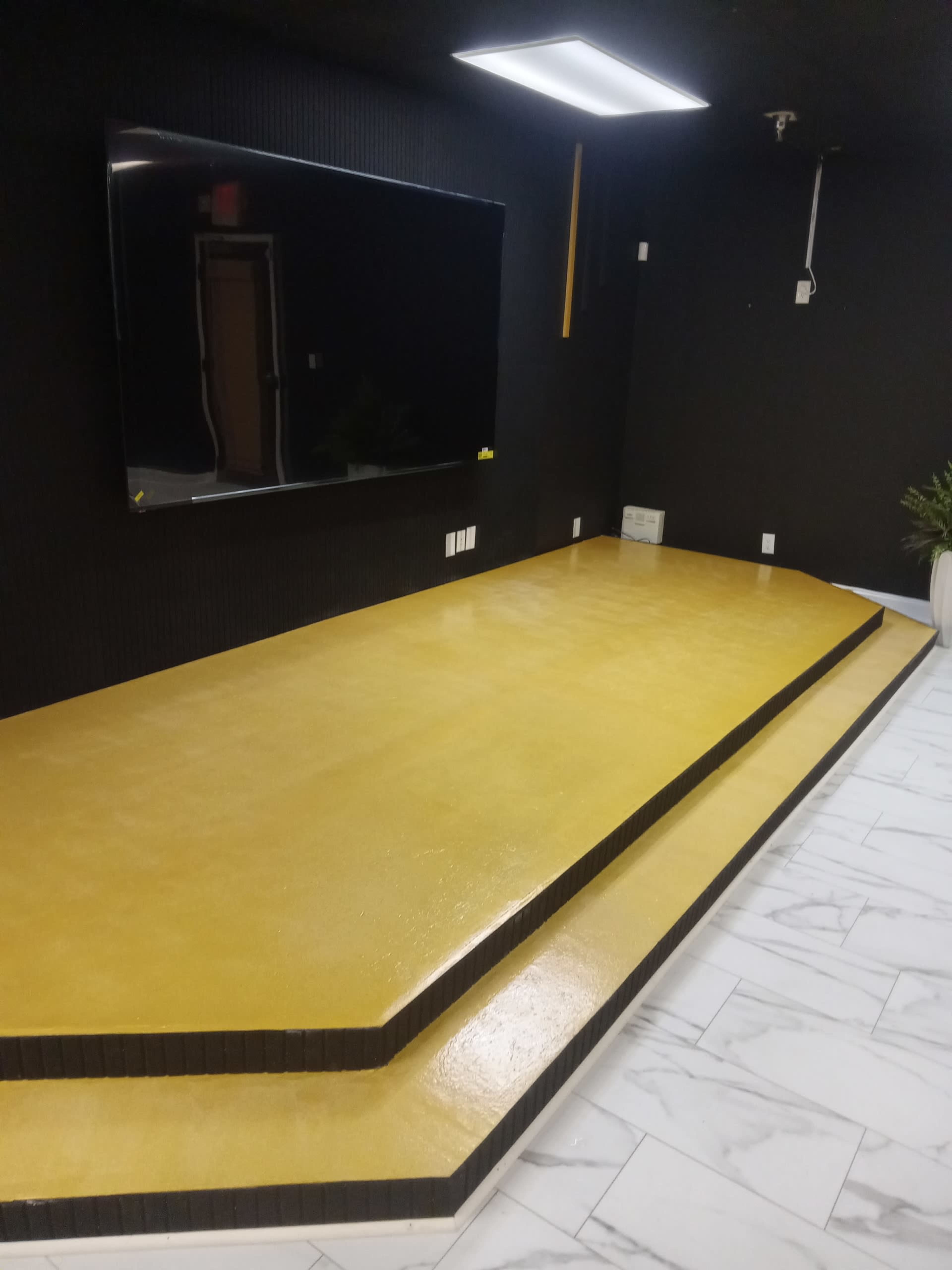 The image shows a stage area with a yellow platform and a large wall-mounted television in a room with black walls and white tiled flooring.