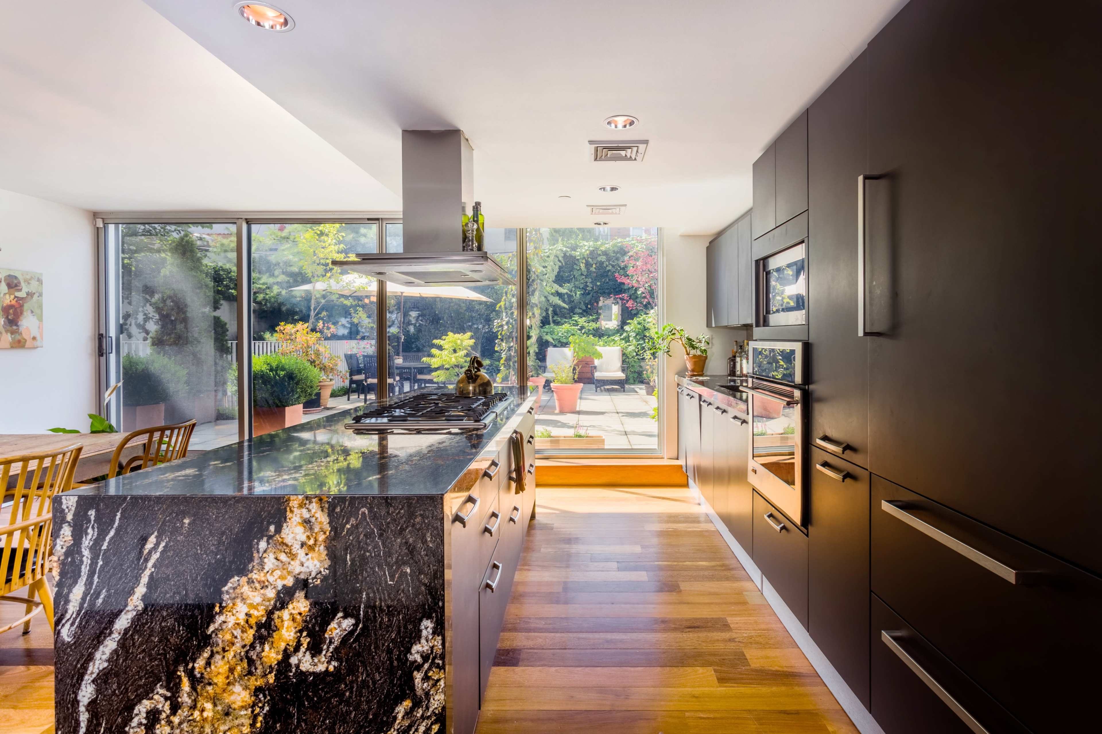 A modern kitchen with a black cabinetry design, a marble countertop, and large windows that open to a garden area.
