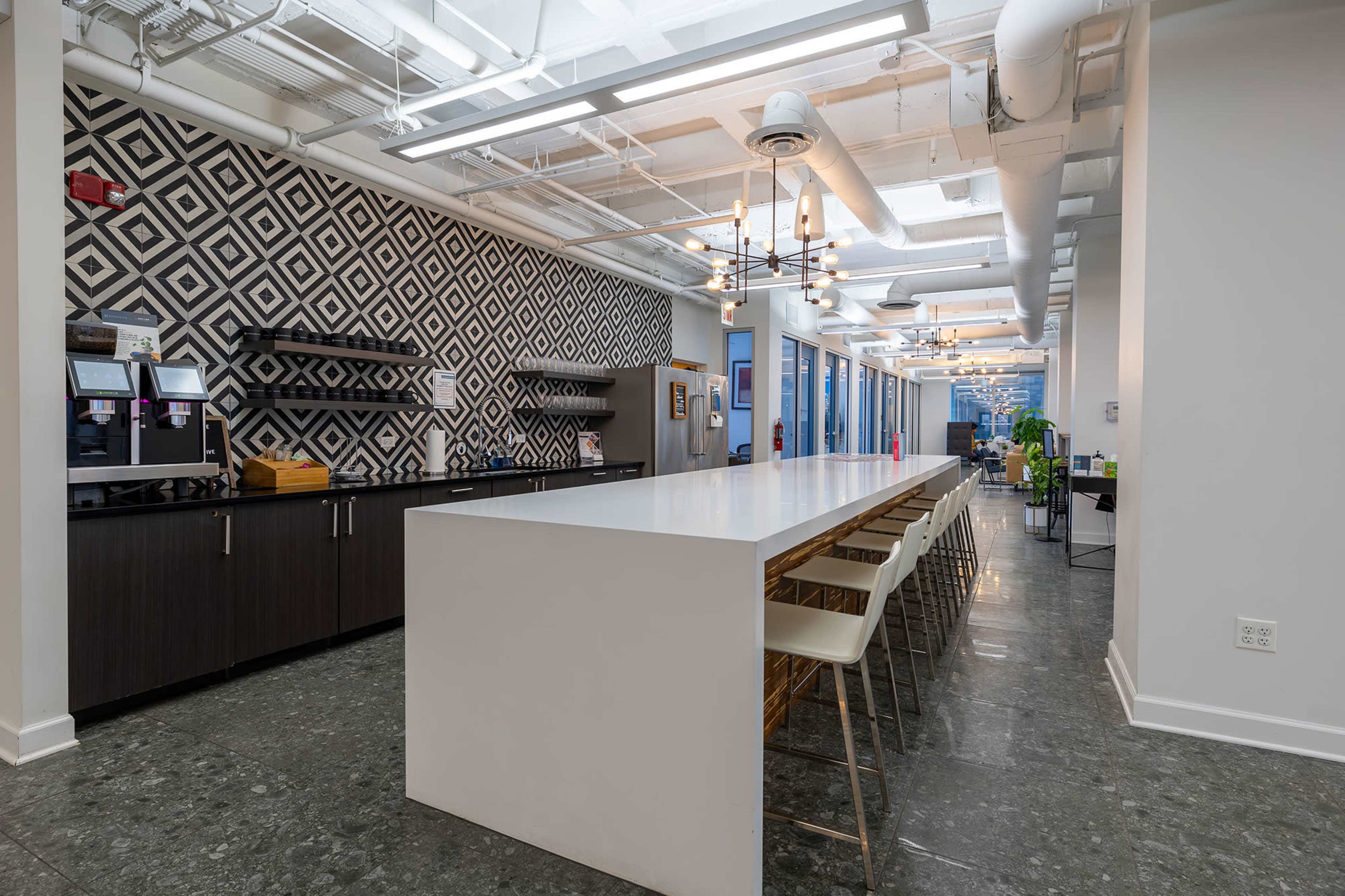 The image shows a modern office kitchen with a long white table and bar seating, featuring a geometric patterned wall and various kitchen appliances.