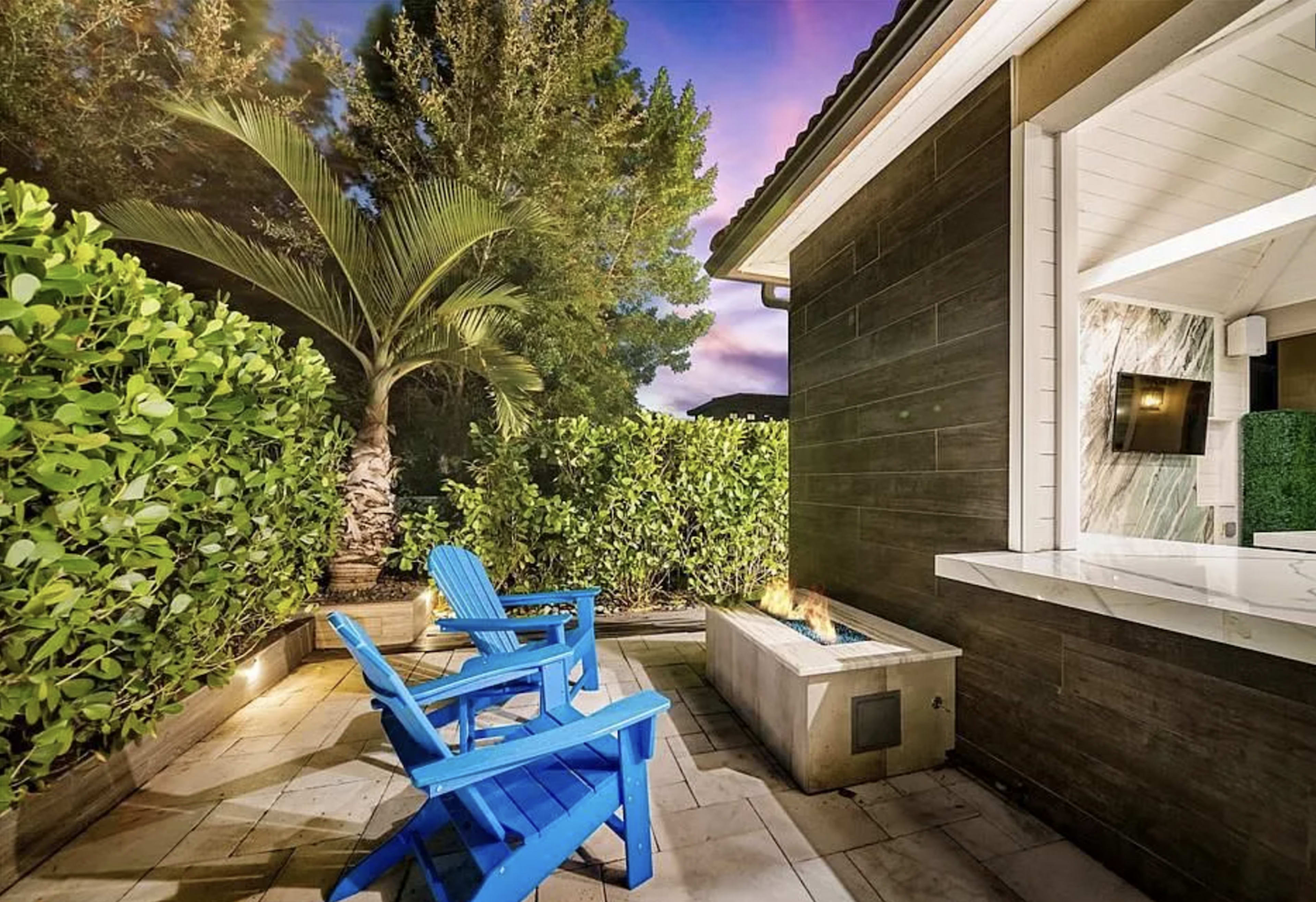 A blue Adirondack chair sits beside a fire pit on a stone patio, surrounded by greenery and a palm tree under a dusk sky.
