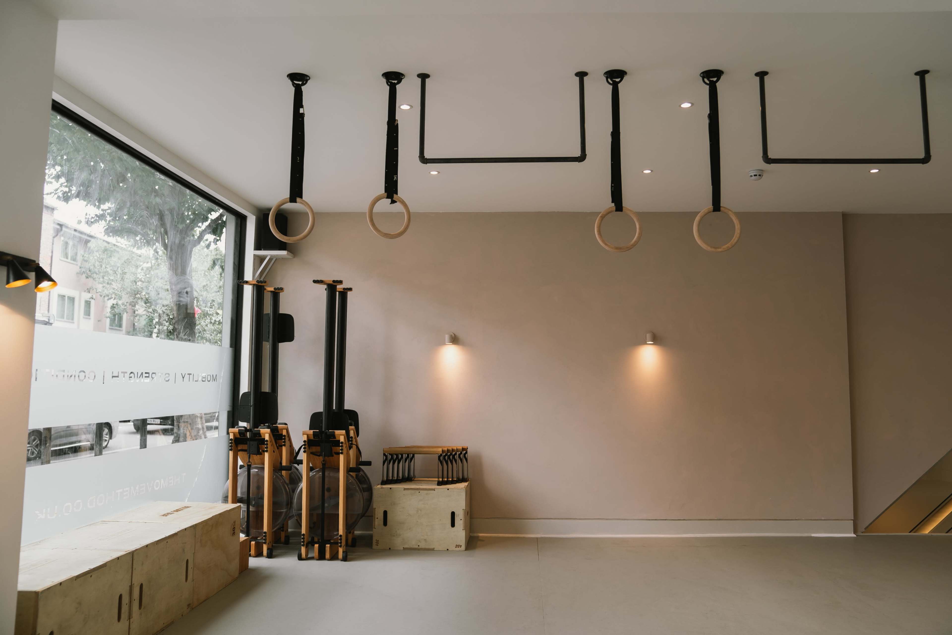 A minimalistic gym space featuring wooden gymnastic rings hanging from the ceiling, along with fitness equipment positioned against the wall.