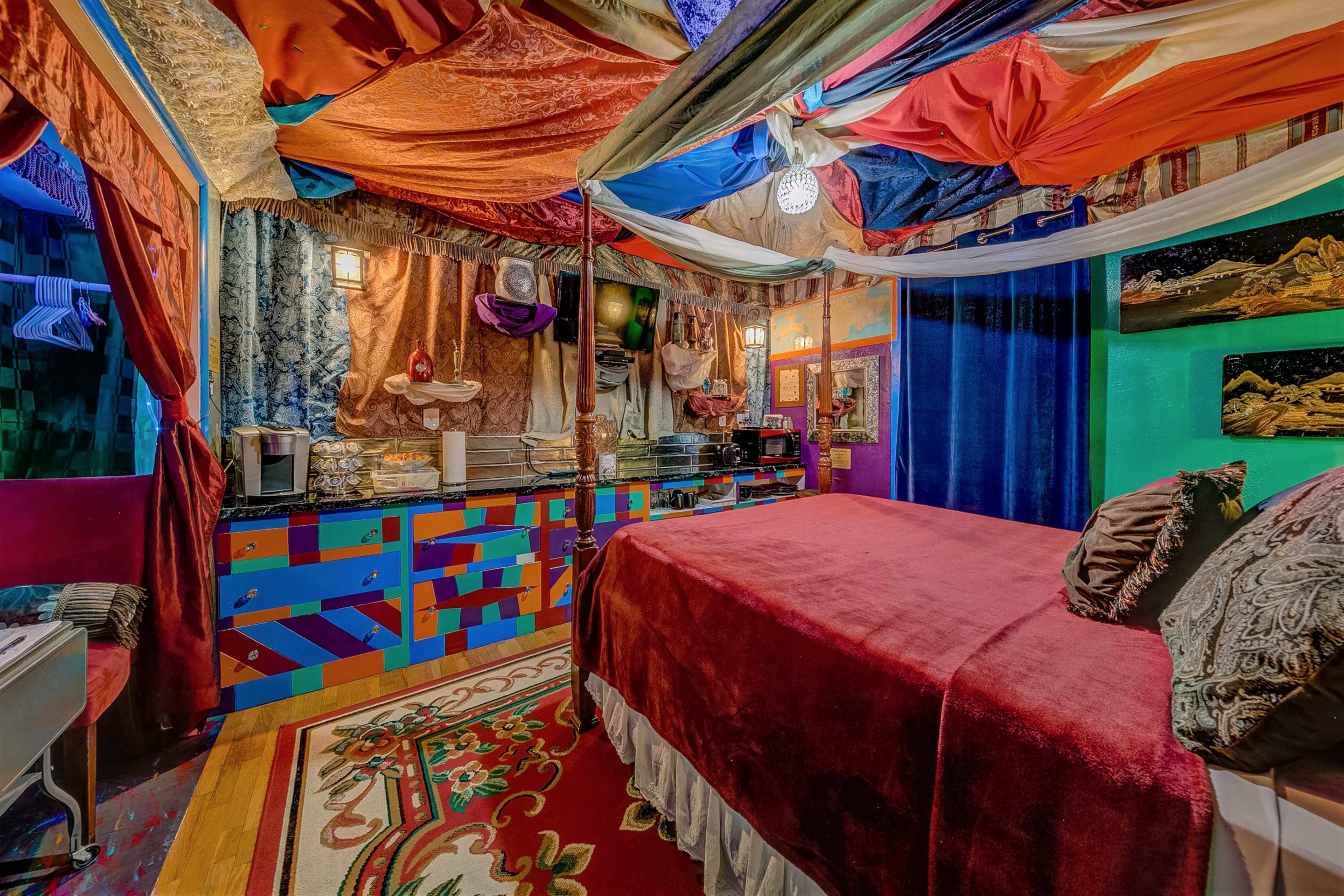 The room features a bed covered with a red blanket surrounded by colorful draped fabric, a decorative rug, and a variety of textiles on the walls.