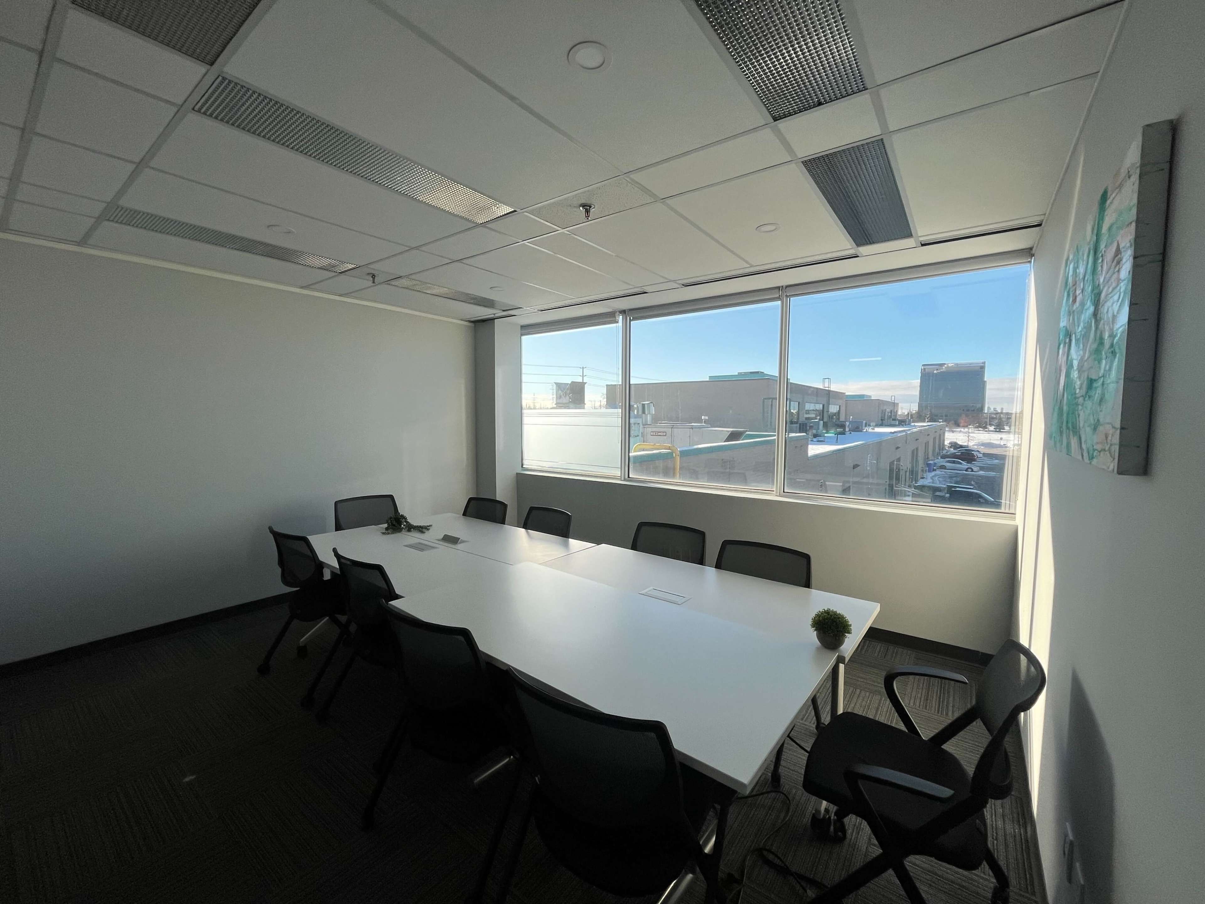 The image shows a well-lit conference room with a long white table surrounded by black chairs, and a large window providing a view of an outdoor urban area.