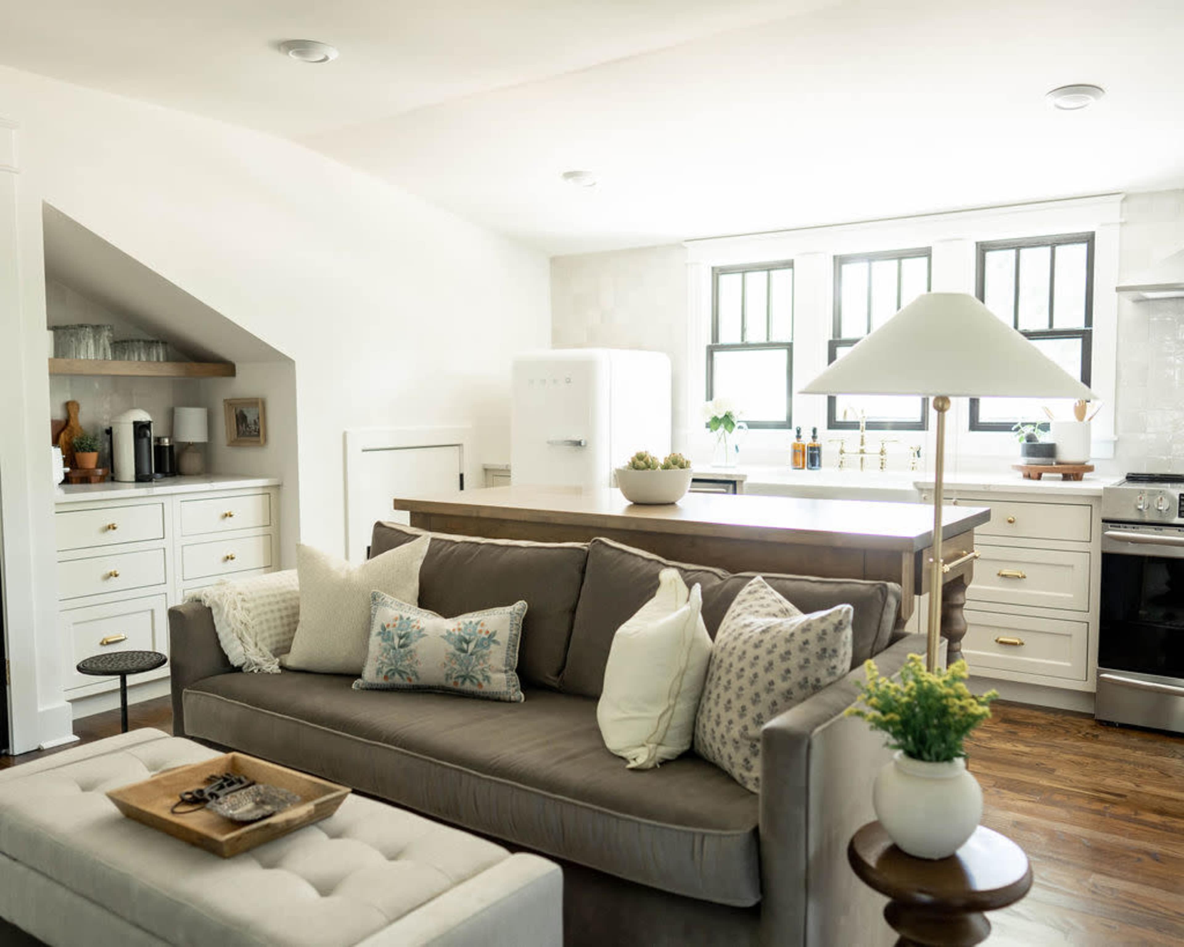 A modern kitchen and living area features a gray sofa with decorative pillows, a large island with bar seating, and a white refrigerator against a backdrop of clean white cabinets and windows.