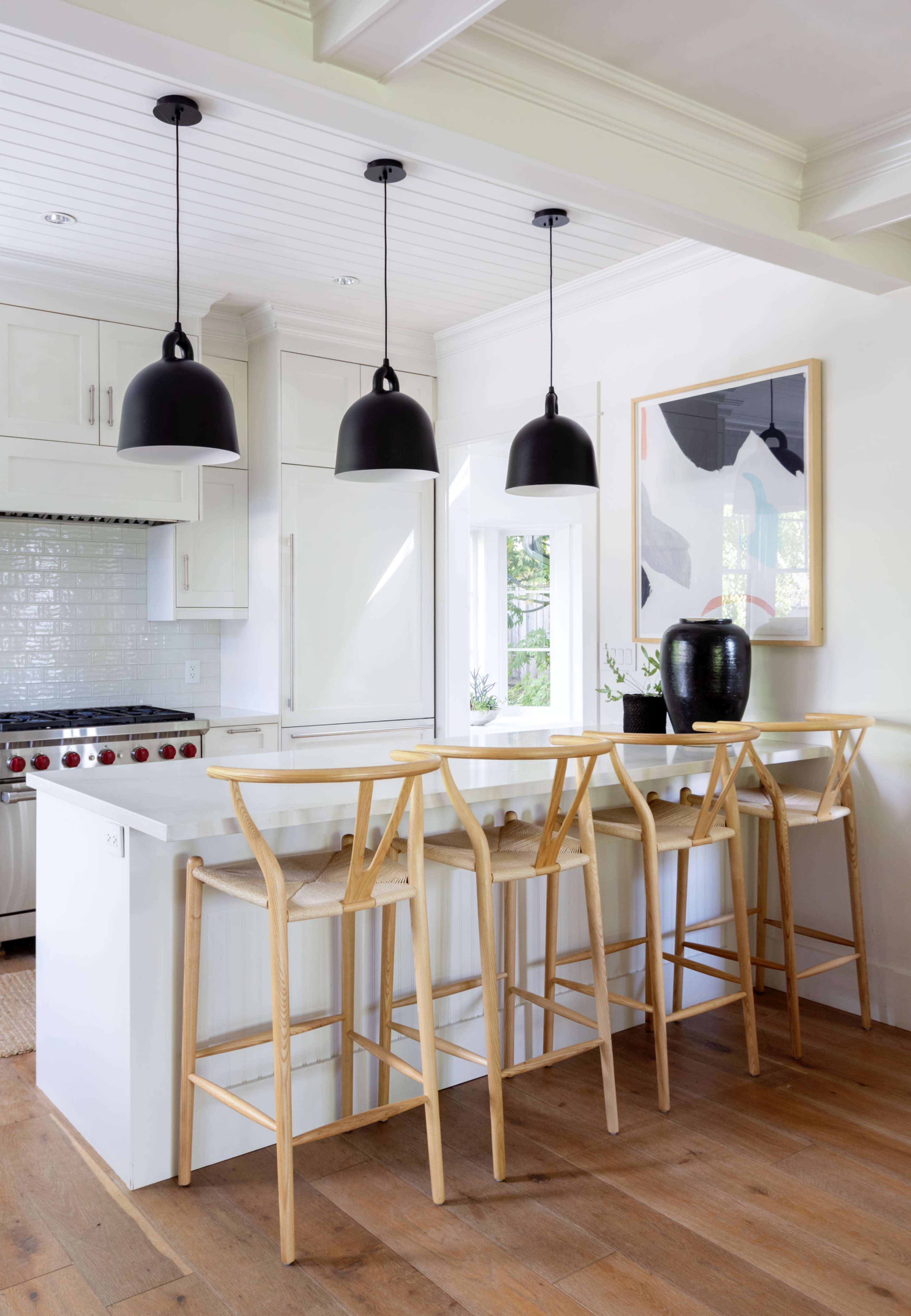 A modern kitchen featuring a white island with four wooden bar stools and three black pendant lights hanging overhead.