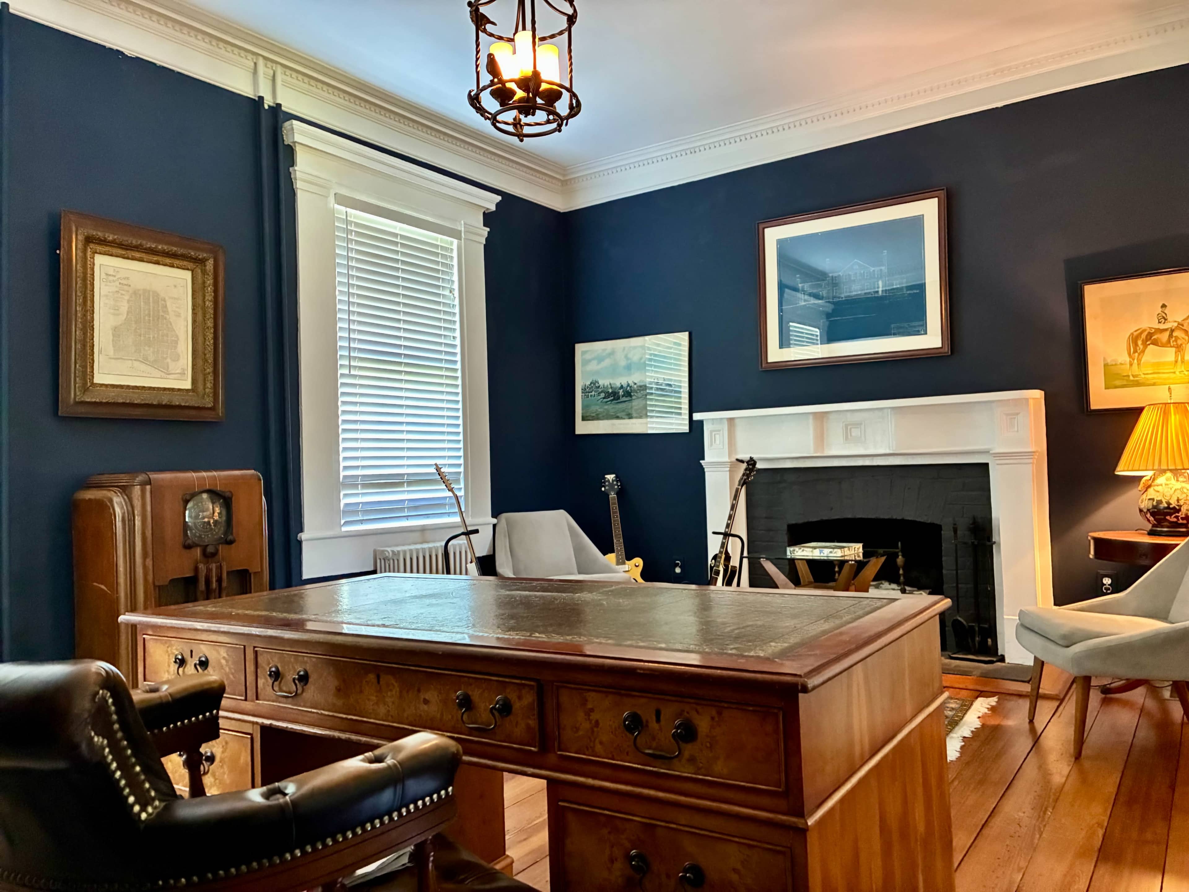 A wooden desk sits in a dark blue room adorned with artwork, a fireplace, and two armchairs.