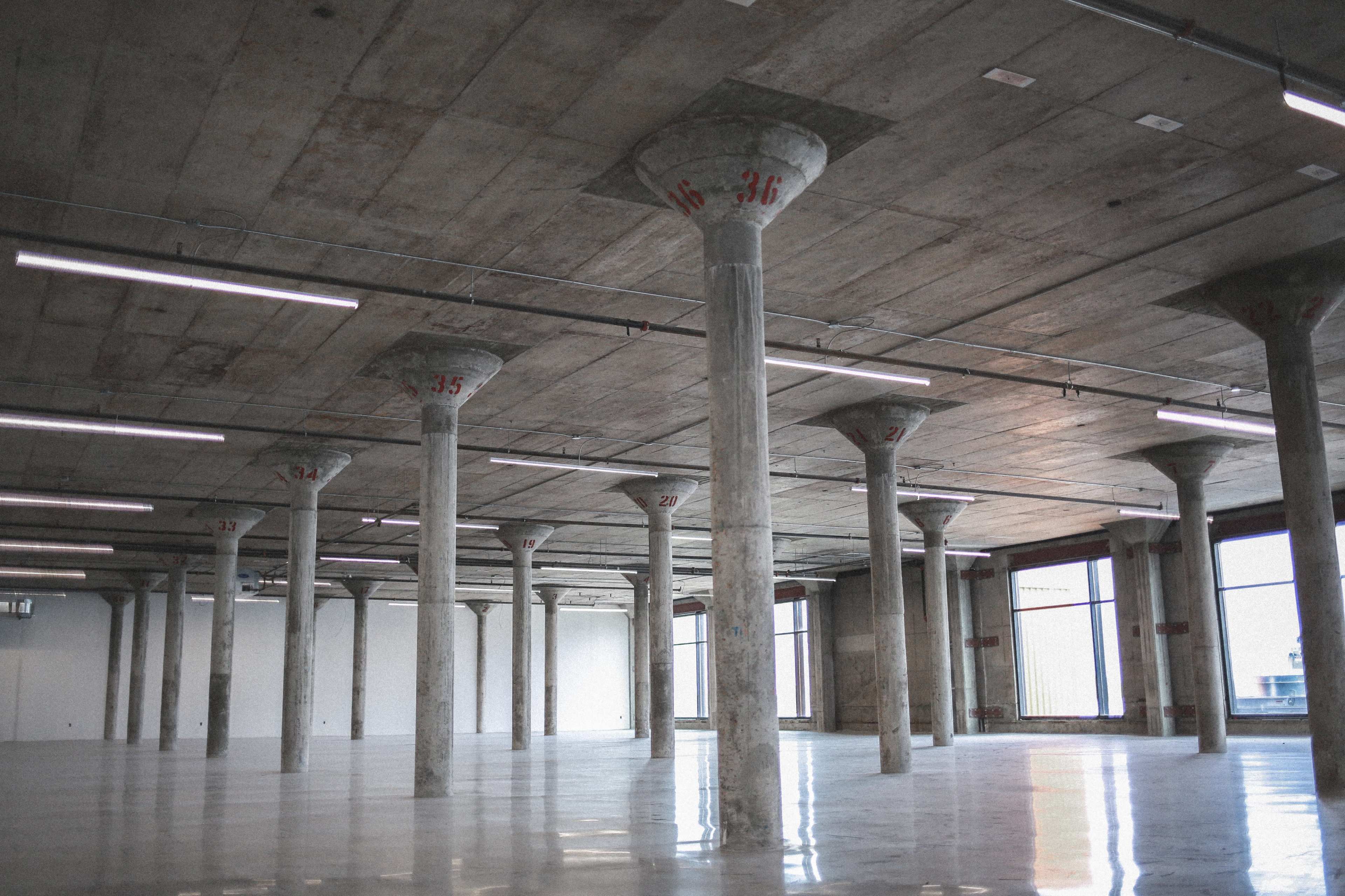 The image shows an empty industrial space with concrete pillars numbered from 22 to 36 and large windows allowing natural light to enter.