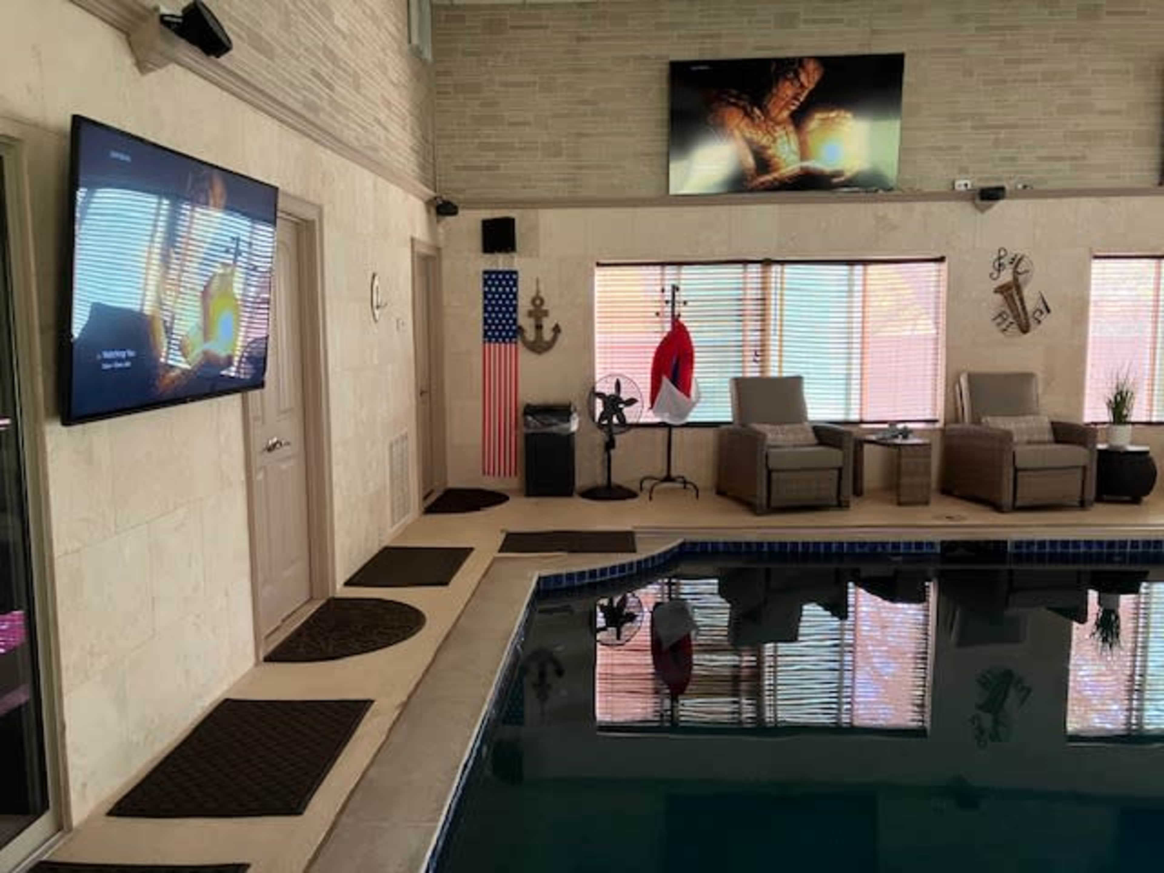 The image shows an indoor swimming pool area featuring a television on a wall, lounge chairs, and decorative elements like an anchor and flags.