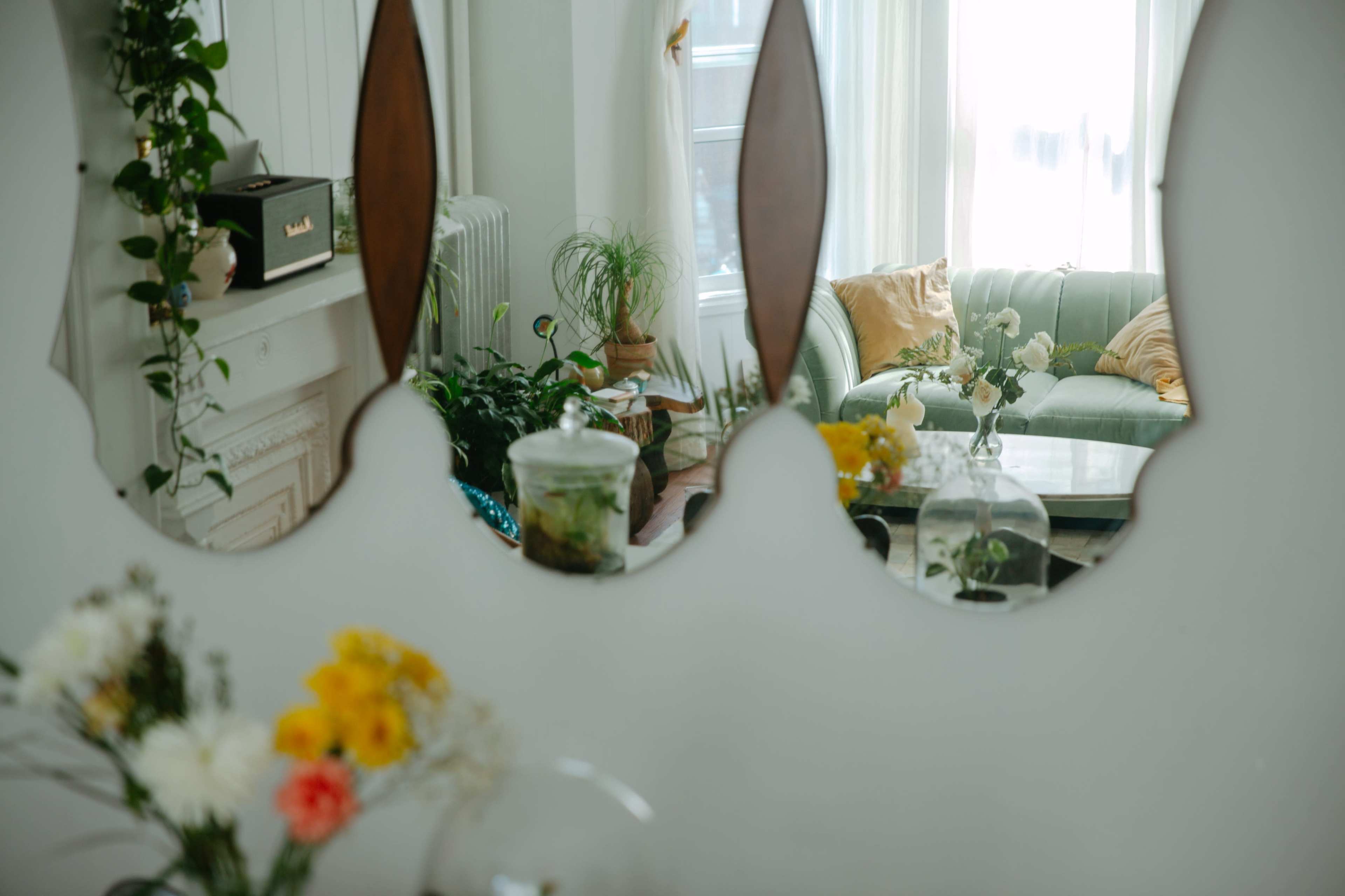 A mirror reflects a bright room with a green sofa, potted plants, and decorative flowers on a table.