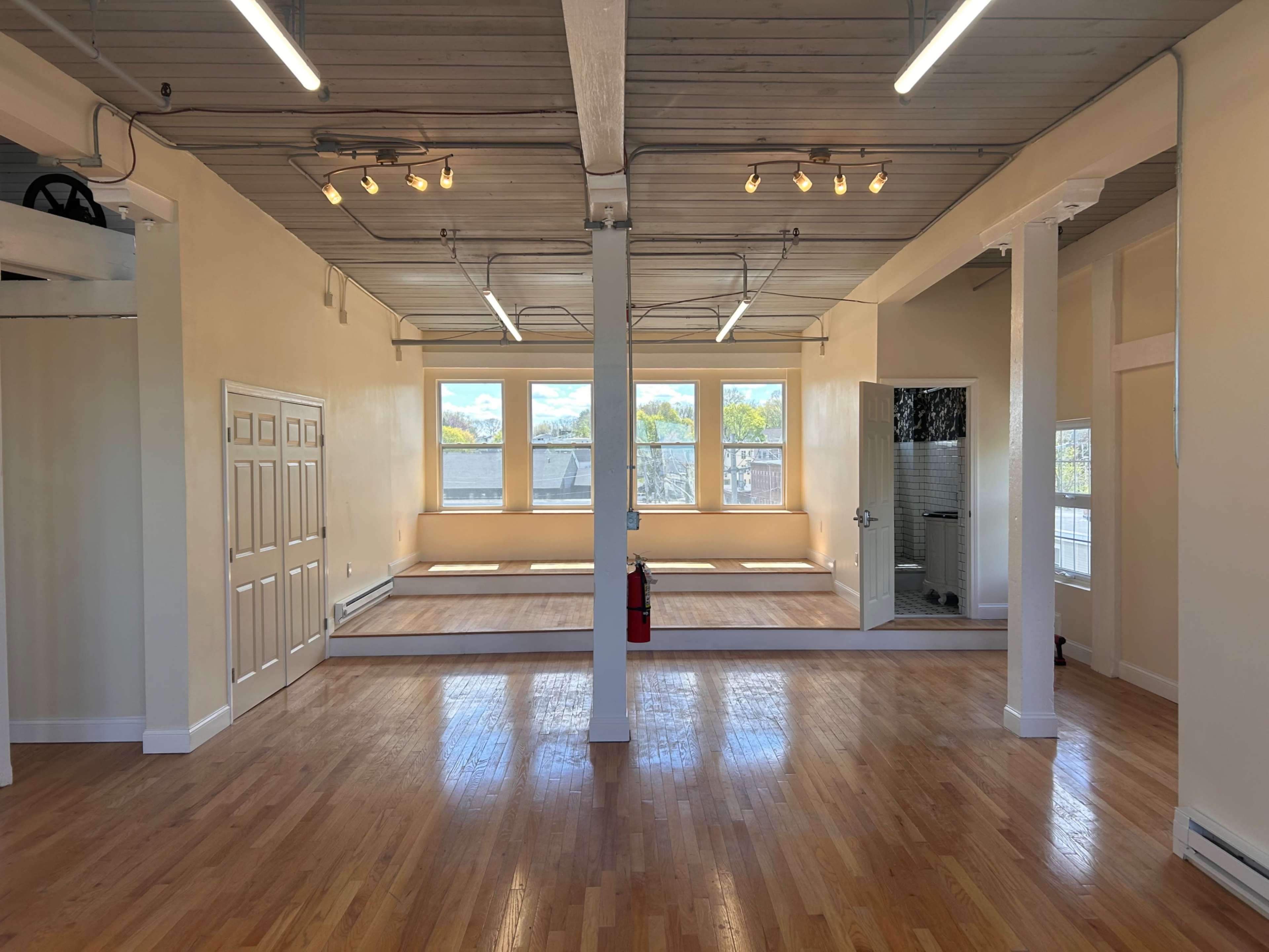 The image shows a spacious, well-lit room with wooden flooring, large windows, and a partitioned area at the center.