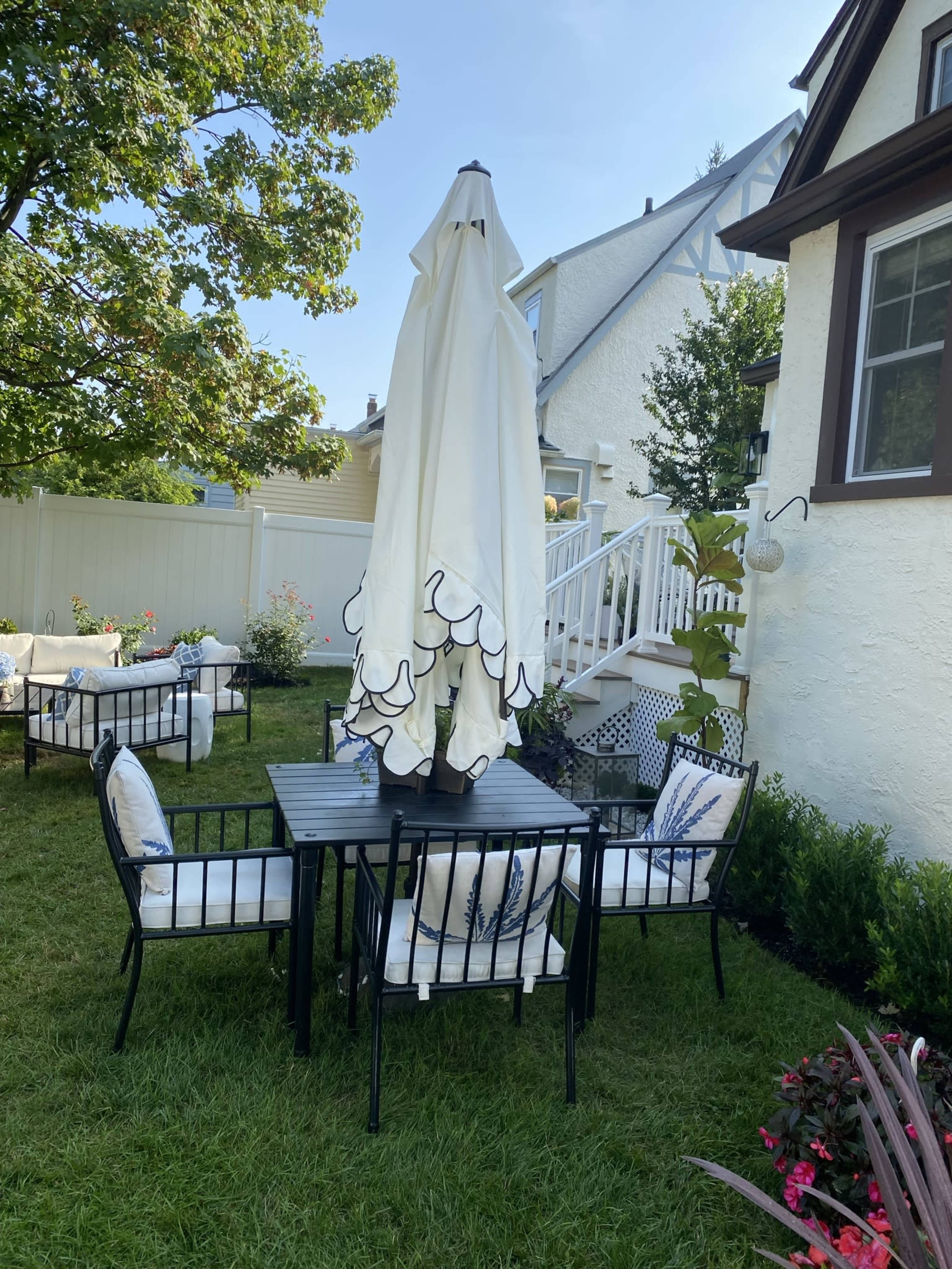 A black metal table with four chairs, covered by a large white umbrella, is set up on a grassy lawn beside a house.