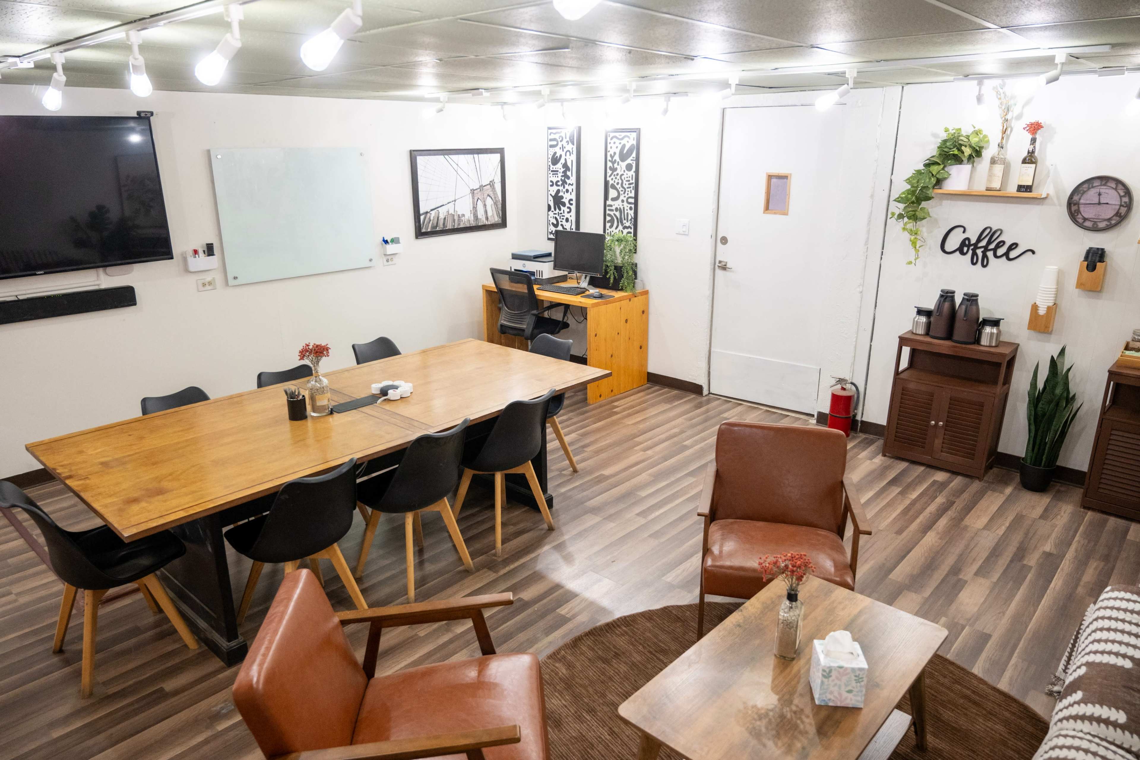The image depicts a cozy meeting room with a large wooden table surrounded by black chairs, an area with a coffee station, and a workspace featuring a computer and wooden storage.