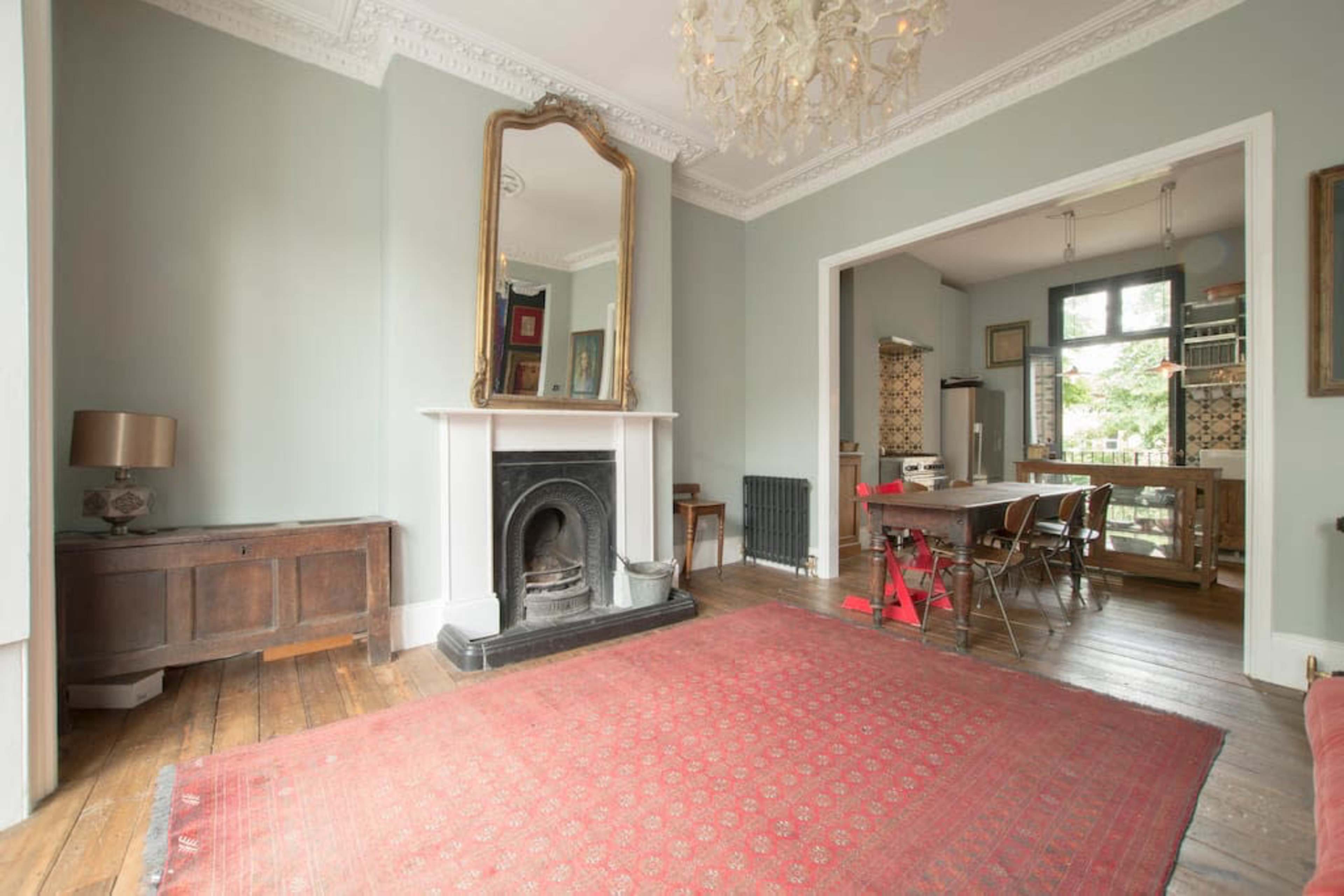 The room features a large mirror above a fireplace, a wooden sideboard, a red area rug, and a dining area with a table and chairs in an adjoining space.