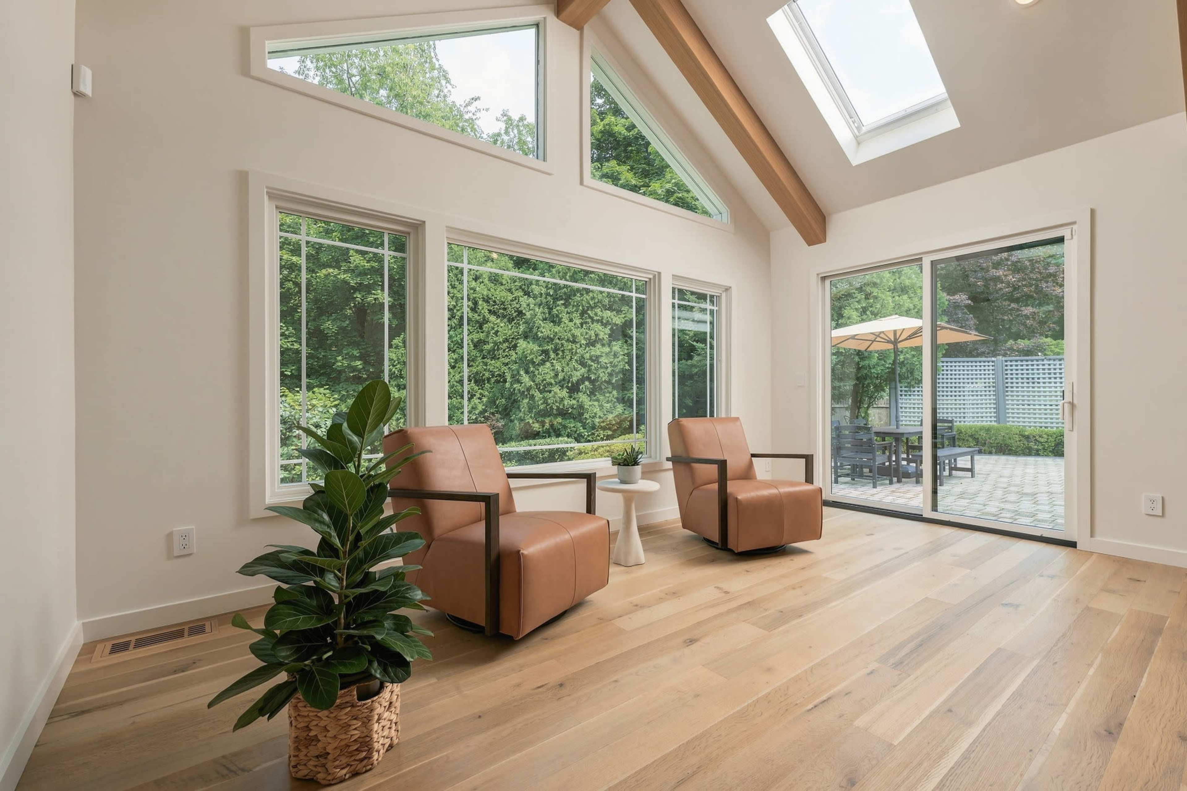 A bright sunroom features two leather armchairs, a small side table, and large windows overlooking a patio with a landscaped garden.